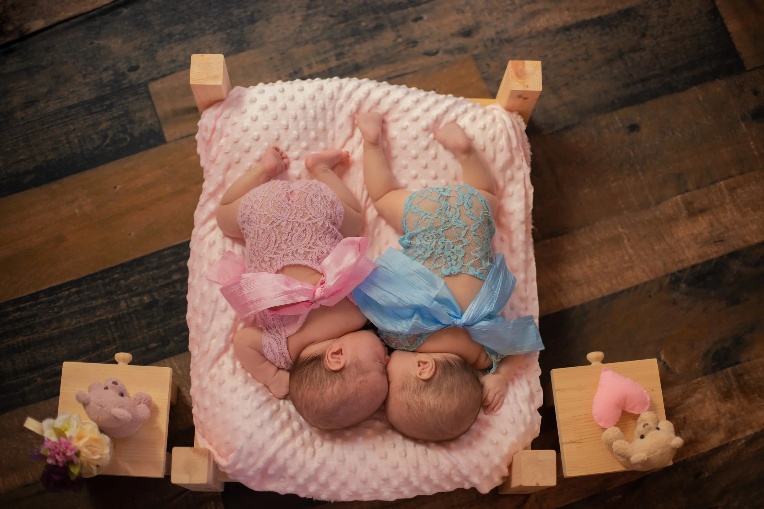 Two identical twin babies lying on pink blanket on a small wooden bed, facing each other, dressed in pink and blue lace outfits with large bows. Two small wooden side tables with plush bear and teddy bear toys and small flower arrangements.
