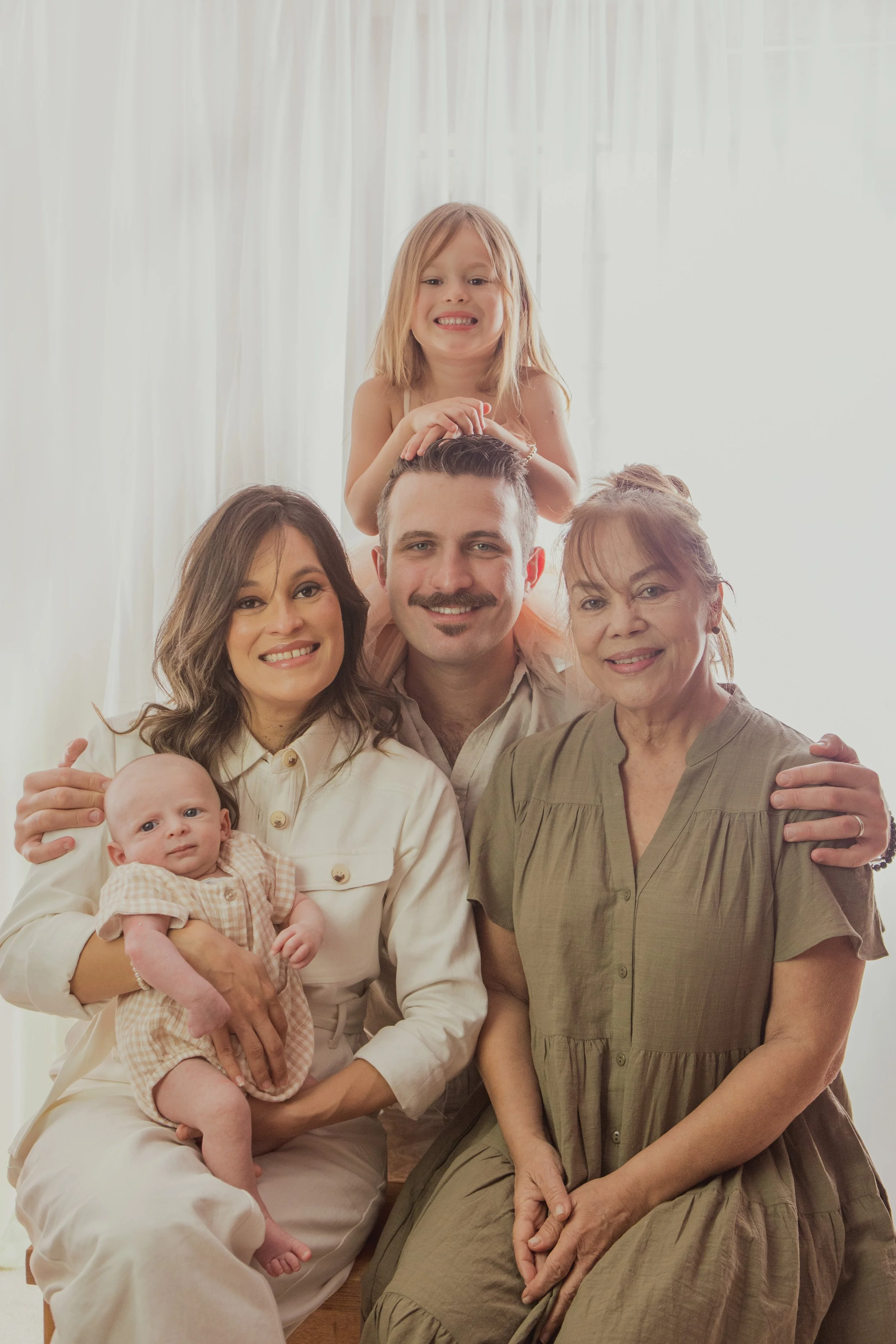 Multi-generational family portrait with three women, a man, and two young girls, all smiling and gathered closely together inside near white curtains.