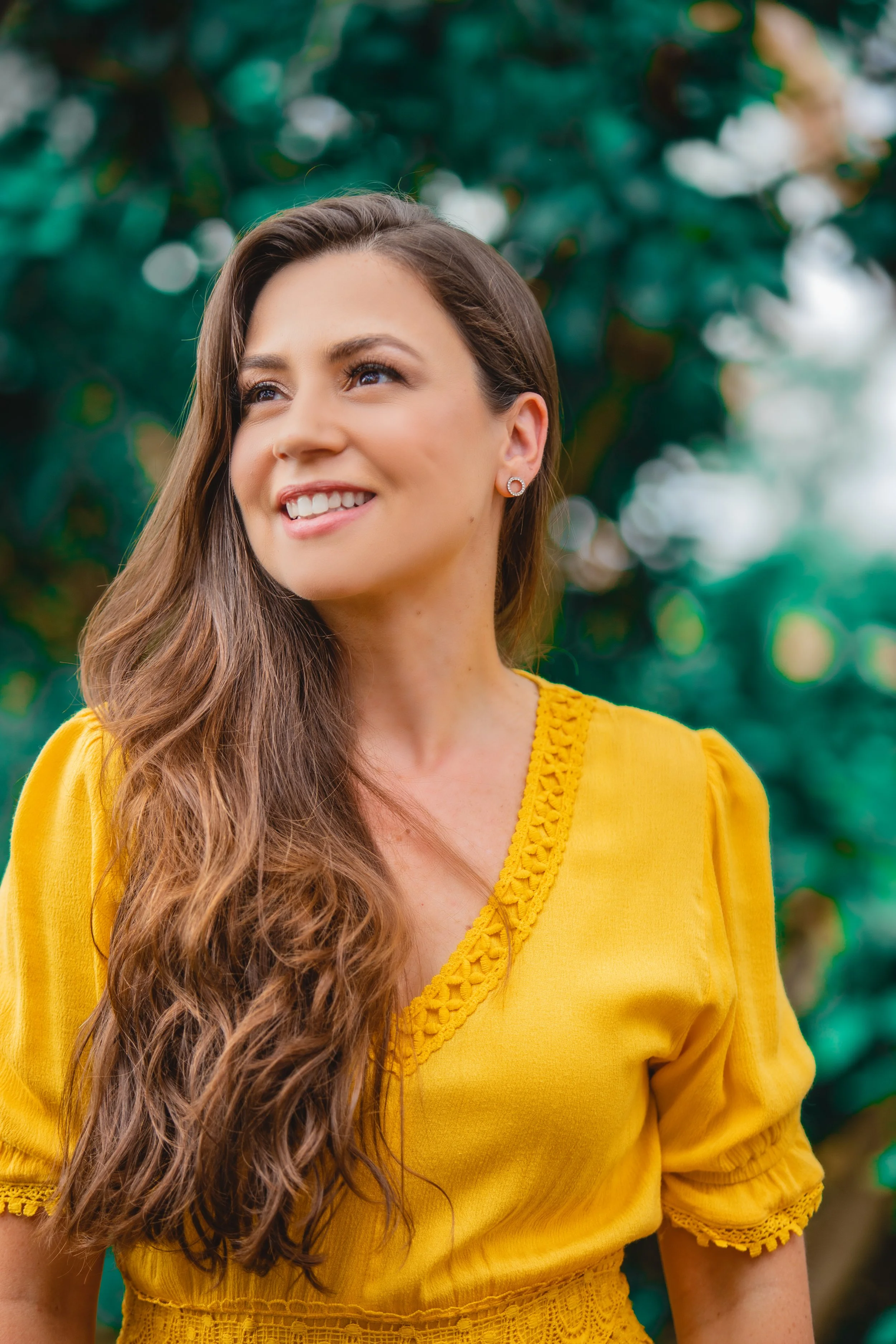 A young woman with long, wavy brown hair wearing a yellow top with lace details, smiling outdoors with a blurred green leafy background.