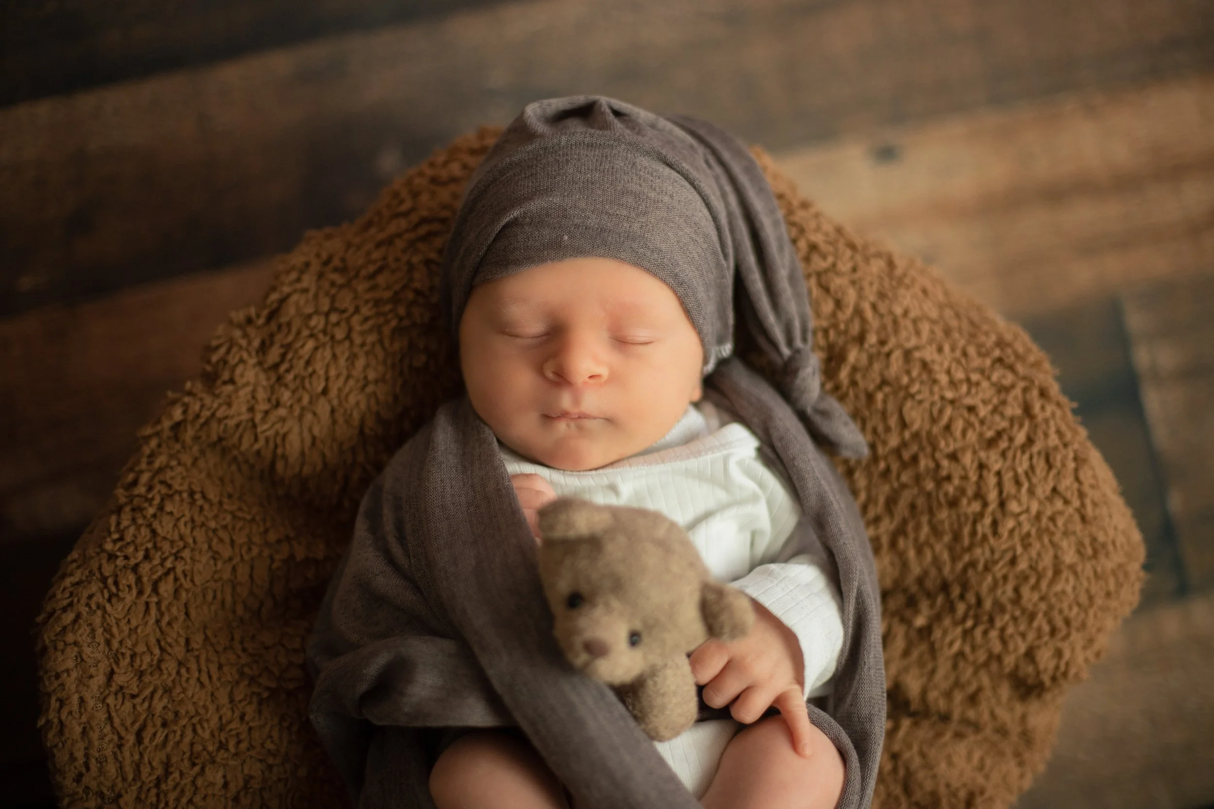 A sleeping baby wearing a gray hat and matching outfit, lying on a brown fuzzy blanket while holding a small teddy bear.