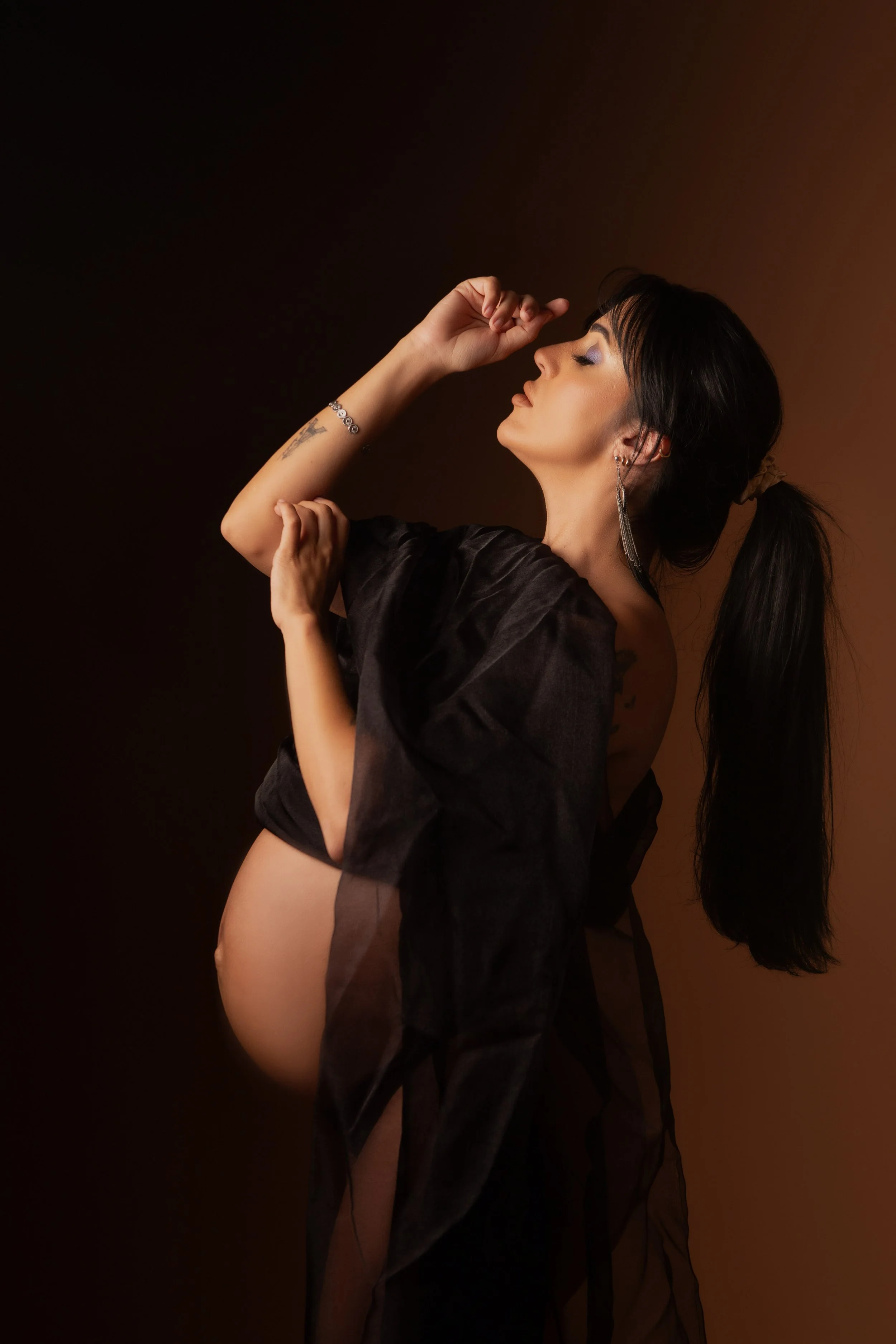 A woman with dark hair styled in a ponytail, wearing large earrings and a black sheer garment, poses with her eyes closed and head tilted back against a dark brown background.