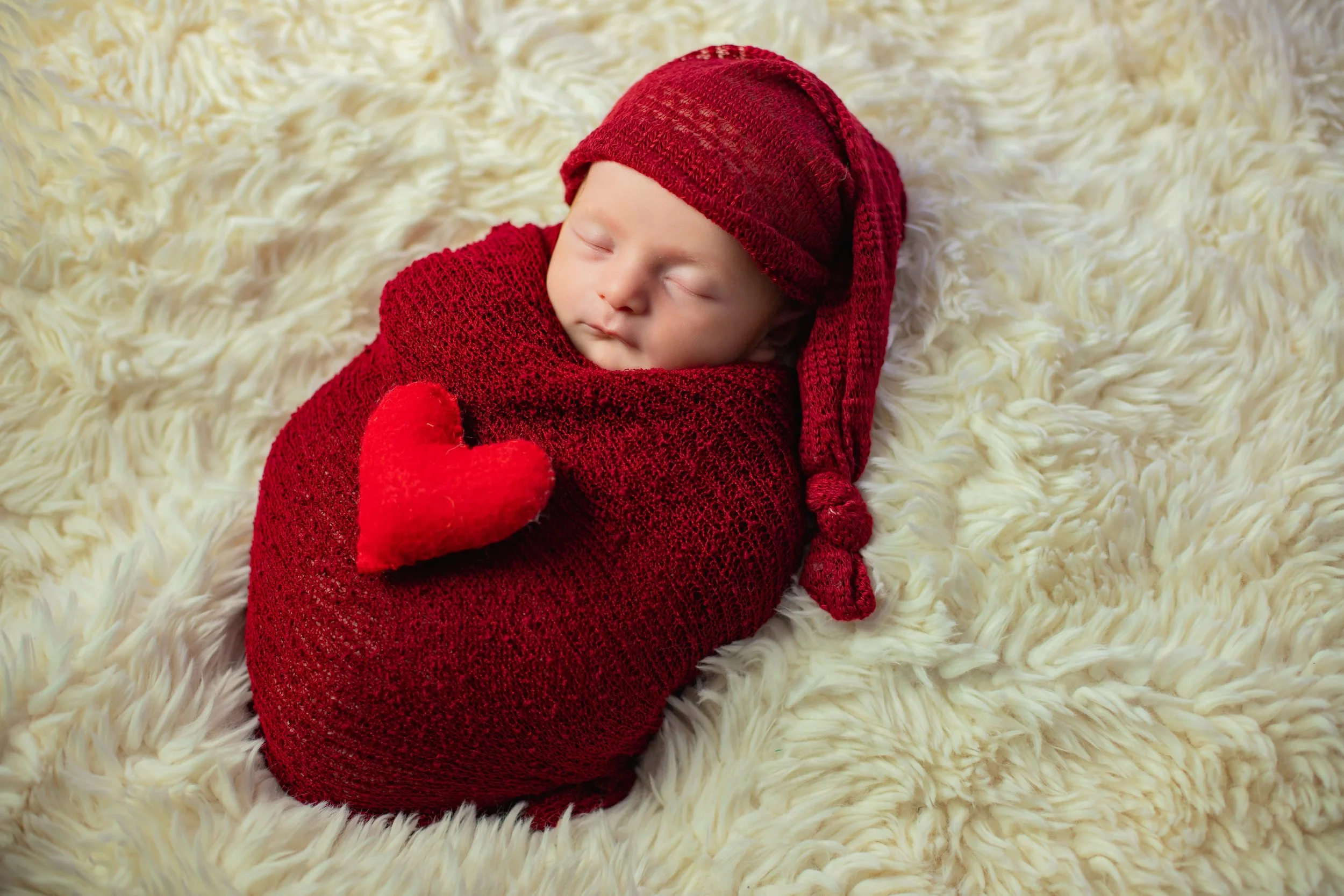 A sleeping baby wrapped in red knitted fabric, holding a red heart, on a fluffy cream-colored blanket, wearing a red hat.
