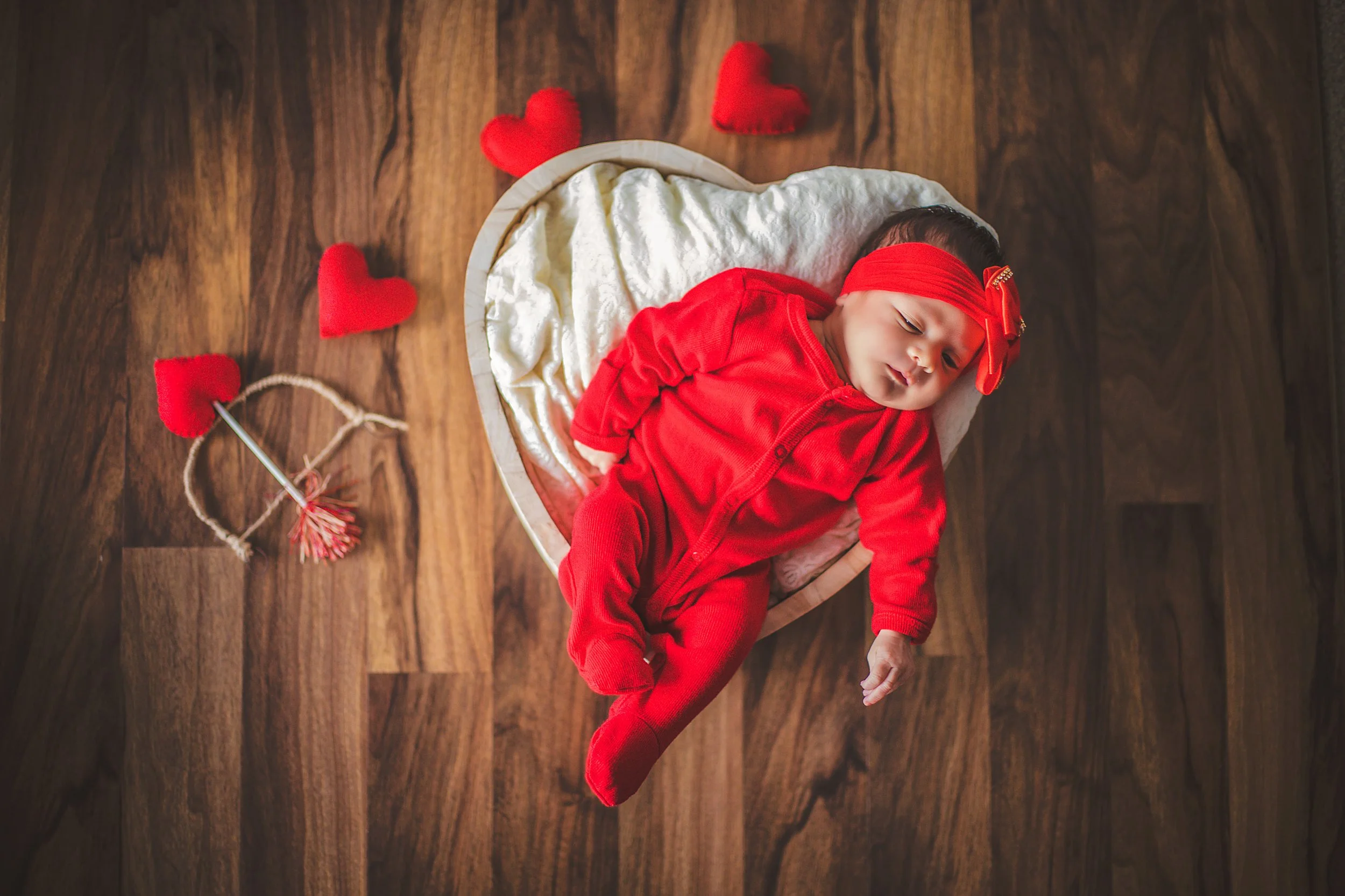 Baby in a red outfit and headband lying in a basket on a wooden floor, surrounded by red heart-shaped decorations.