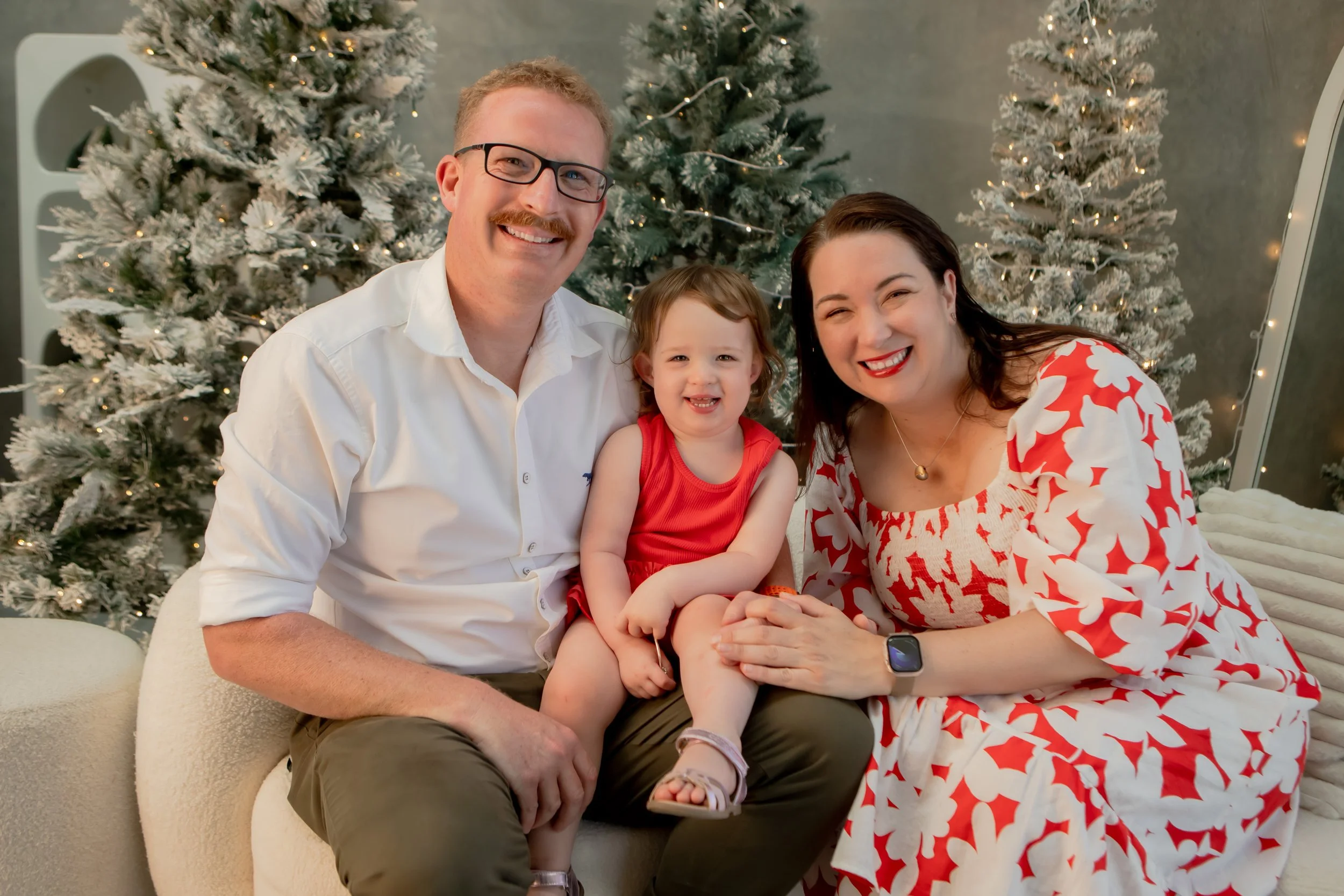 A family of three, a man, a woman, and a young girl, smiling and sitting together in front of decorated Christmas trees with lights. The man is wearing glasses and a white shirt, the woman is in a white and red patterned dress, and the girl is in a r