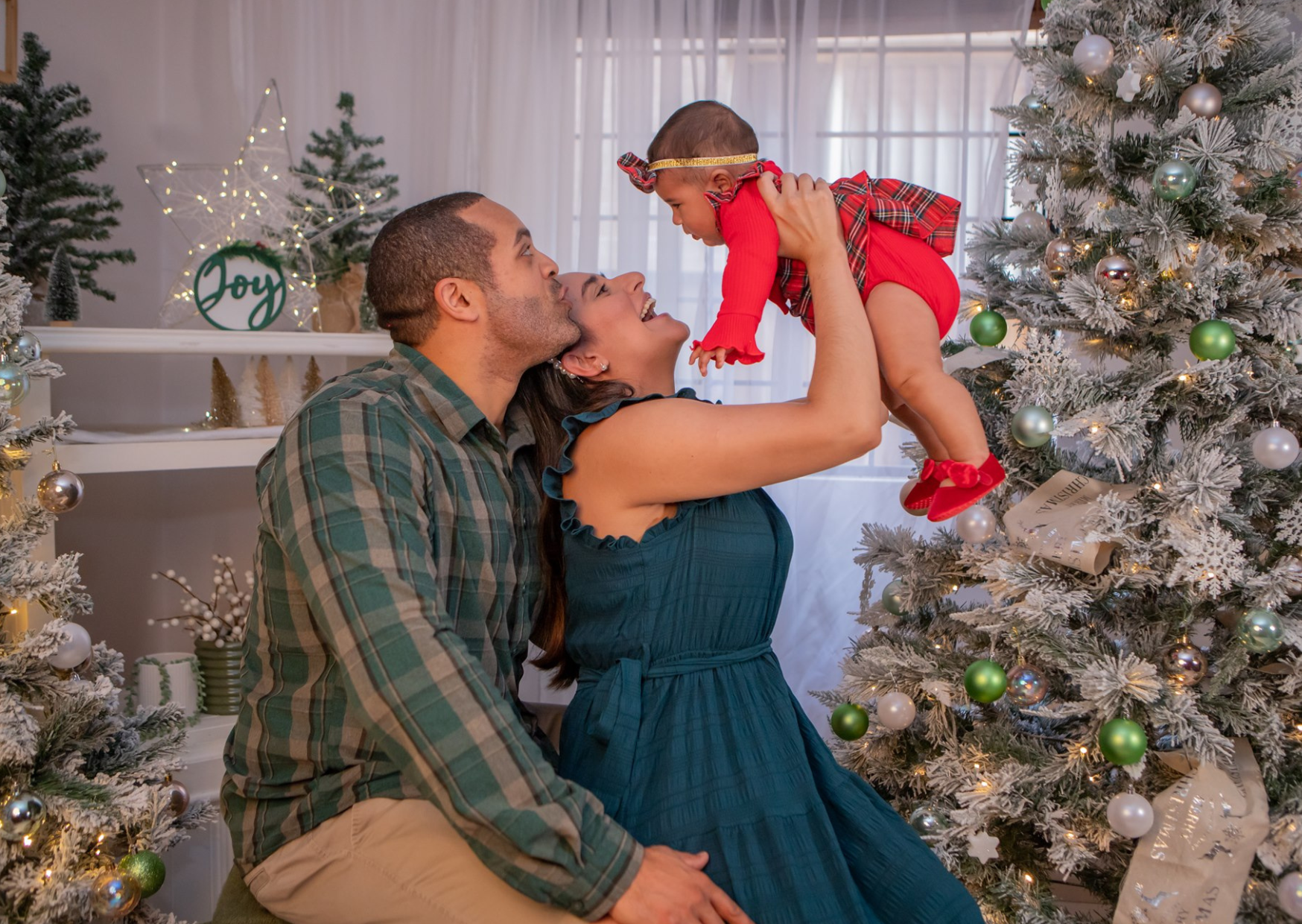 A family of three celebrating Christmas near a decorated Christmas tree, with the mother holding her young daughter up in the air as they smile at each other.