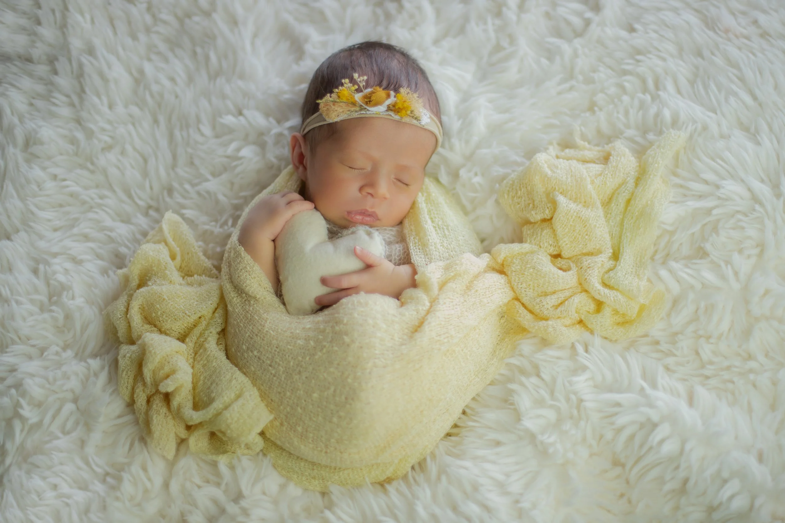 Adorable newborn baby girl sleeping on a fluffy white blanket, wearing a yellow headband with daisies and a yellow knitted blanket wrapped around her.