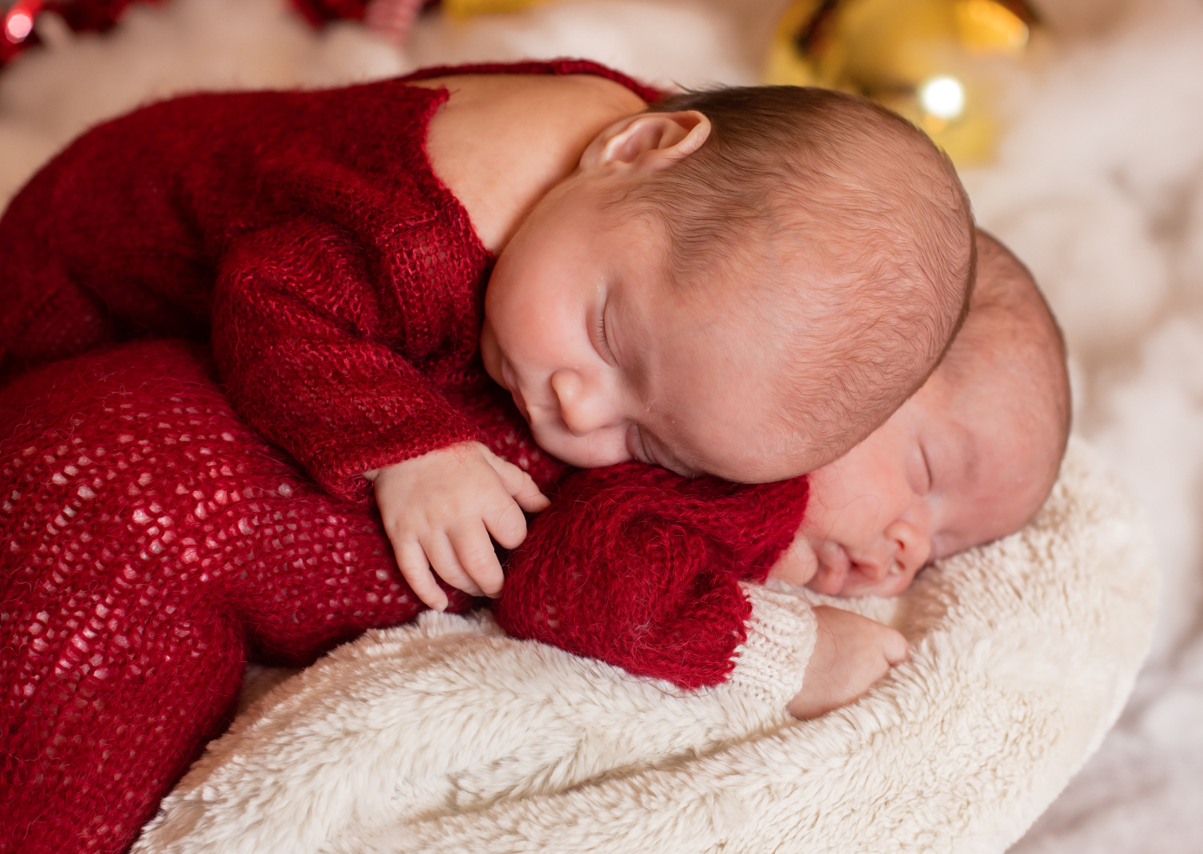 Two babies in red sweaters asleep on a soft white blanket with a blurred Christmas tree in the background.