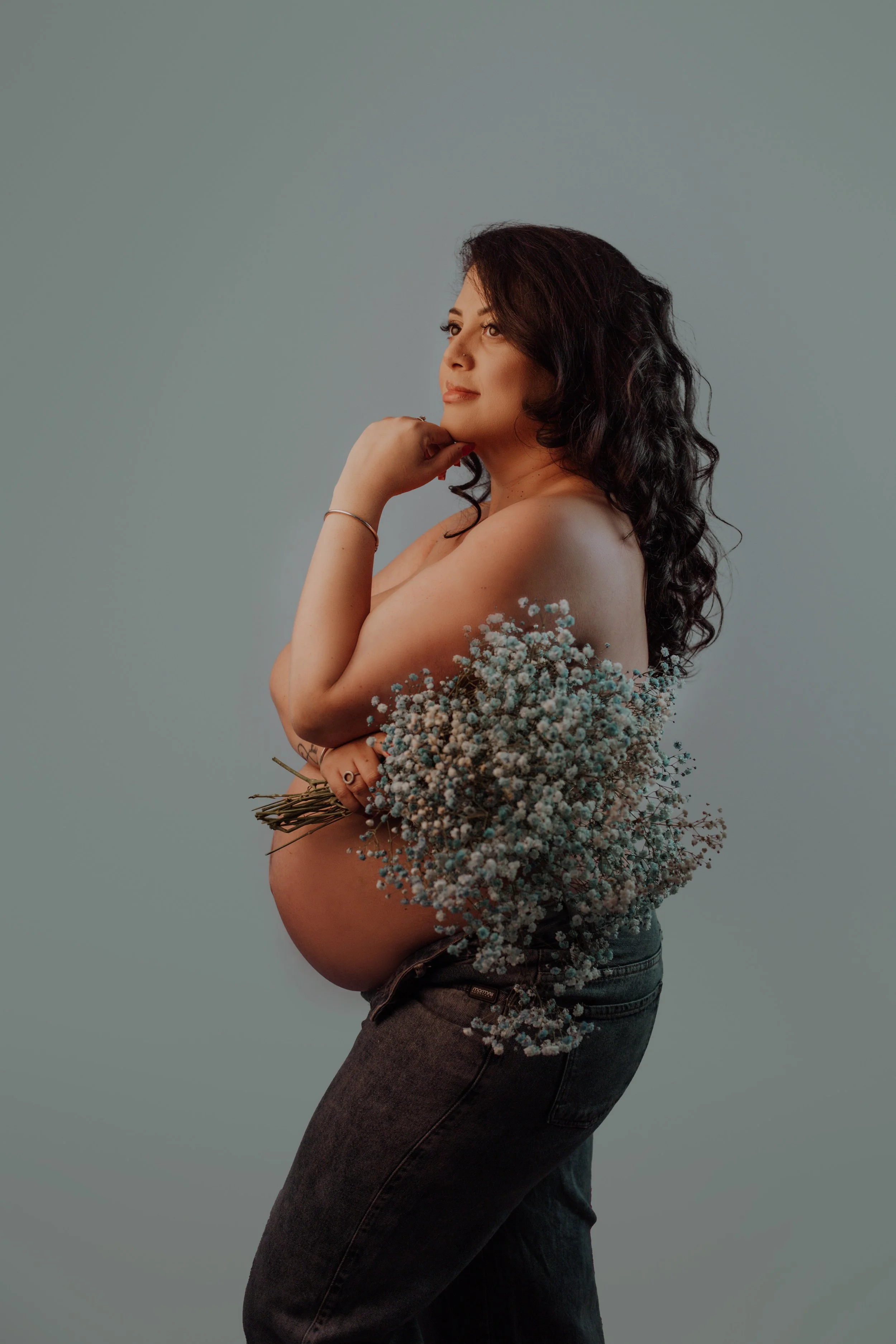 Pregnant woman holding a bouquet of baby's breath flowers, topless with her hand resting on her chin, against a plain background.