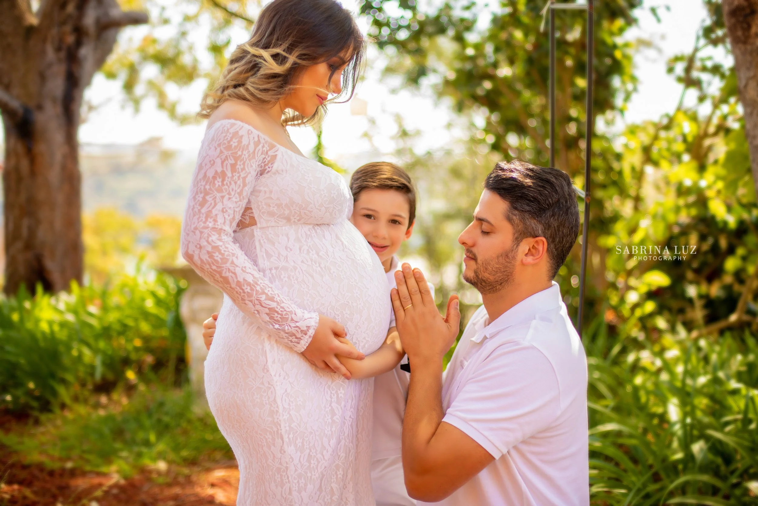 A man kneeling with eyes closed, touching his forehead to a pregnant woman's belly while a young boy stands behind her, smiling, in a garden setting.