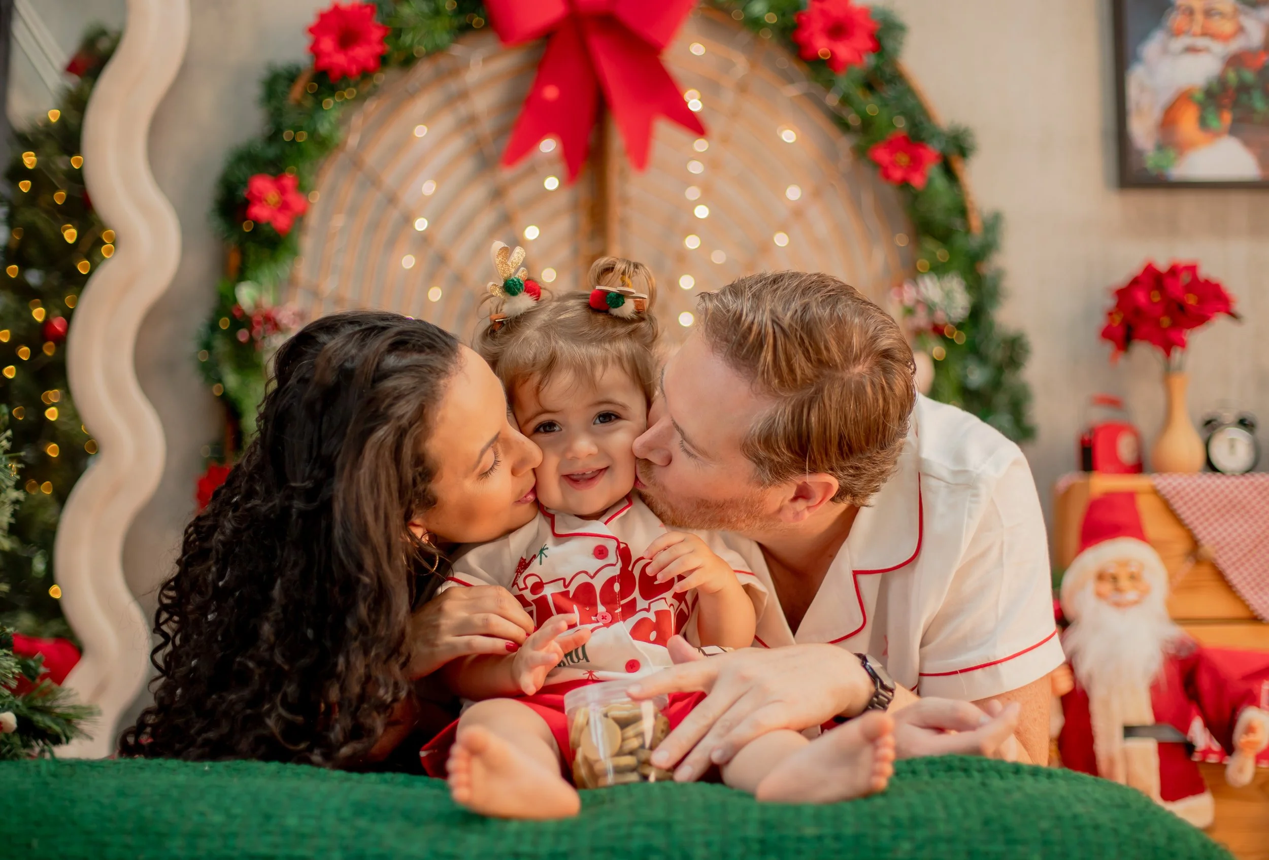 A family of three, a woman, a man, and a young girl, happily sharing a moment during Christmas time with Christmas decorations, including a decorated tree, poinsettias, and Santa Claus figure, in the background.