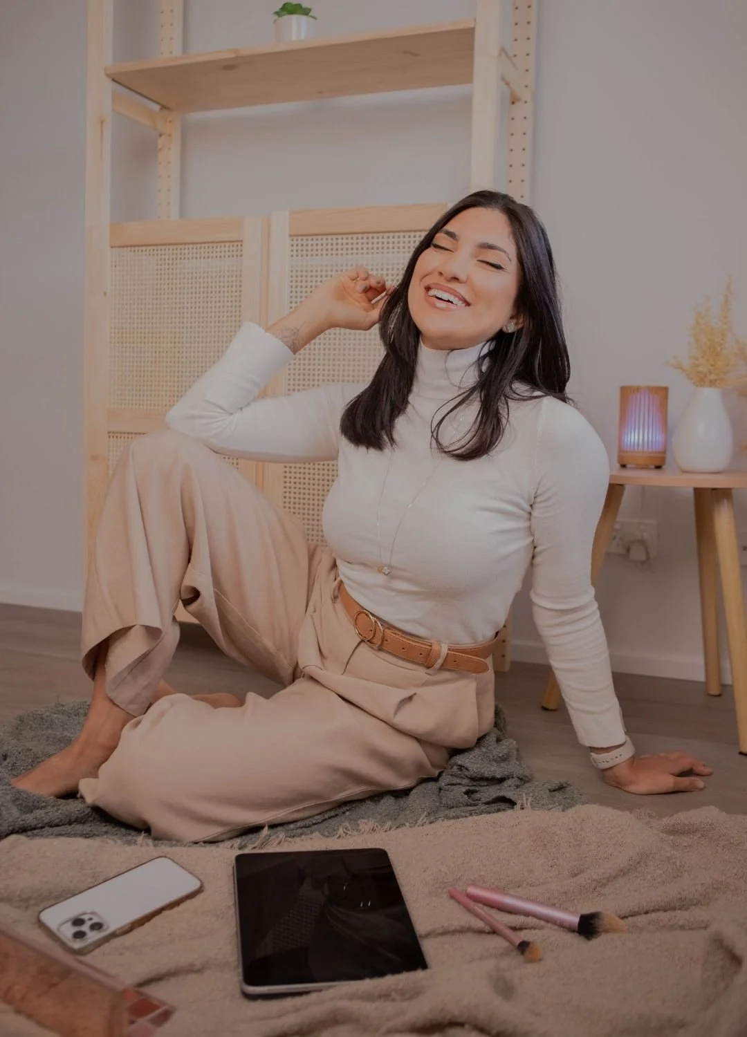 A woman with dark hair is sitting on a soft blanket on the floor, smiling with her eyes closed, wearing a white turtleneck and beige wide-leg pants. She is leaning back on her left hand with her right hand near her head. There are makeup brushes, a s