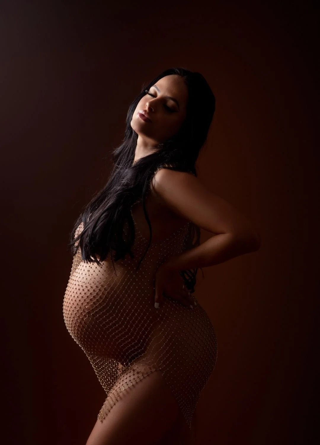 A woman with long dark hair posing against a dark background, wearing a sheer, beaded dress with one hand on her hip and eyes closed.
