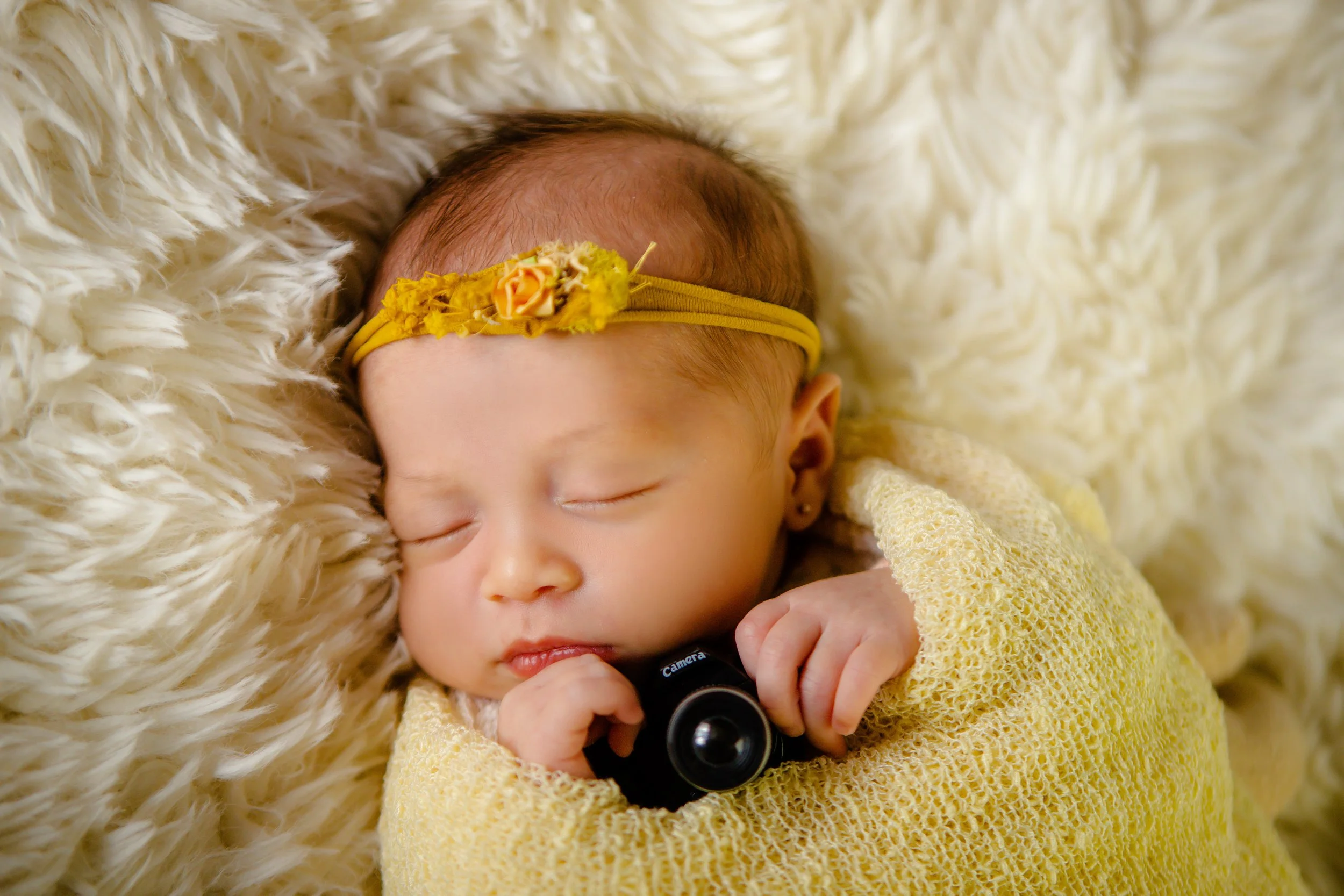 A sleeping baby girl with a yellow headband and a yellow knit sweater lying on a fluffy cream-colored blanket, holding a black camera toy.