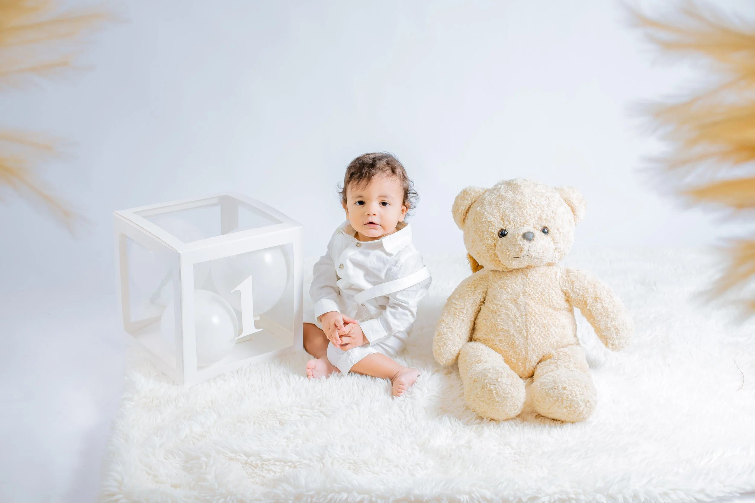 Baby sitting on a white fluffy rug next to a large teddy bear and a cube with white balloons and the number one inside.