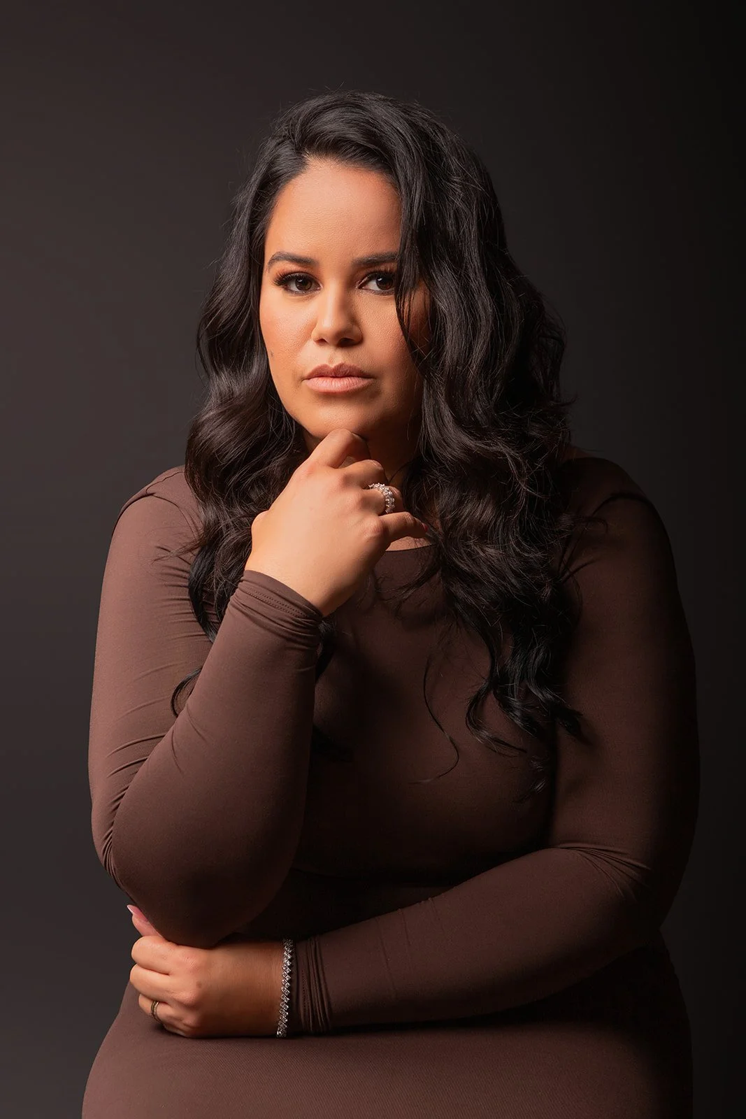 A woman with dark, wavy hair posing against a dark background, resting her chin on her hand, wearing a brown long-sleeve dress and jewelry.