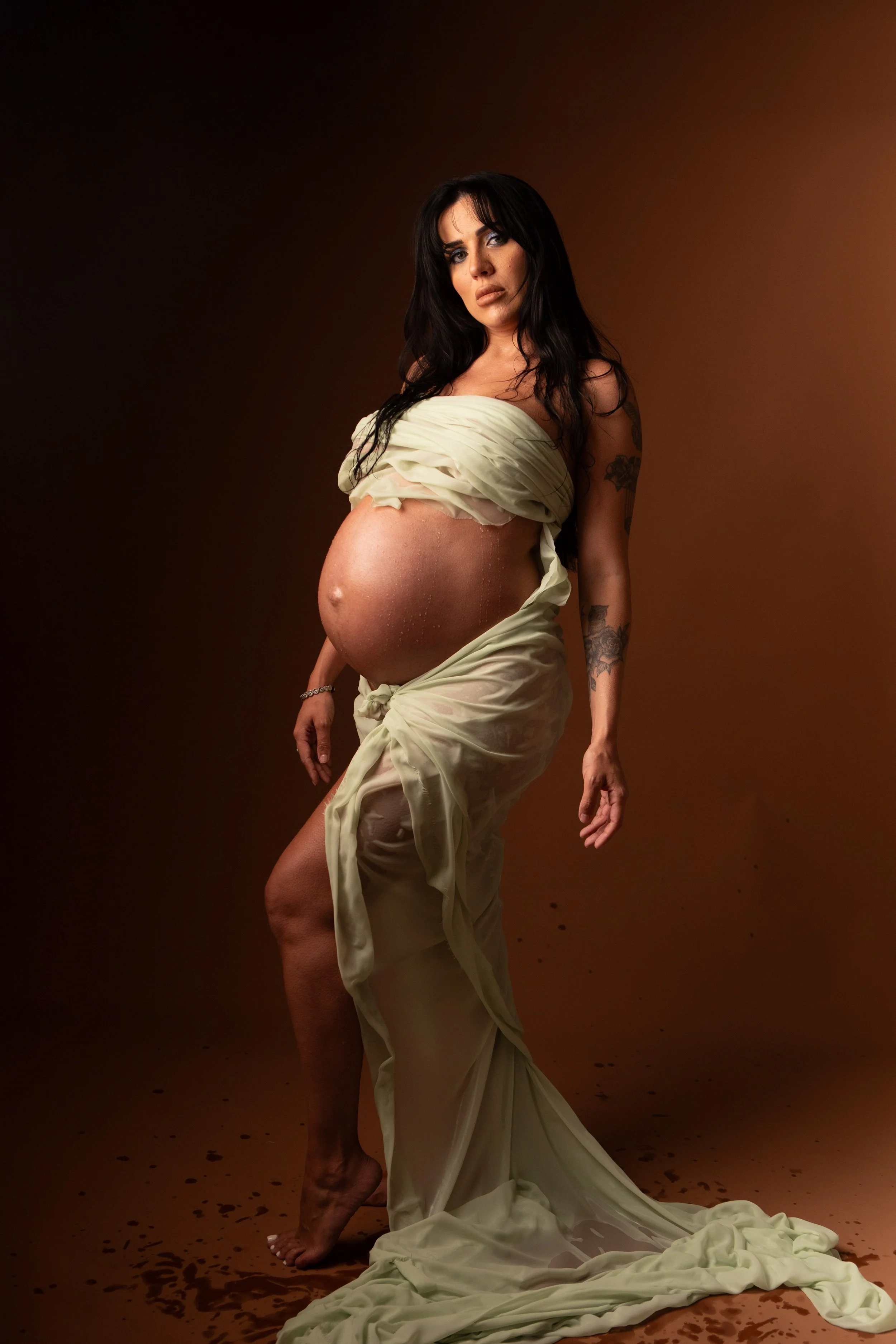 Pregnant woman with long dark hair wearing a sheer, off-white fabric draped around her, standing barefoot on a brown floor with a dark brown background.