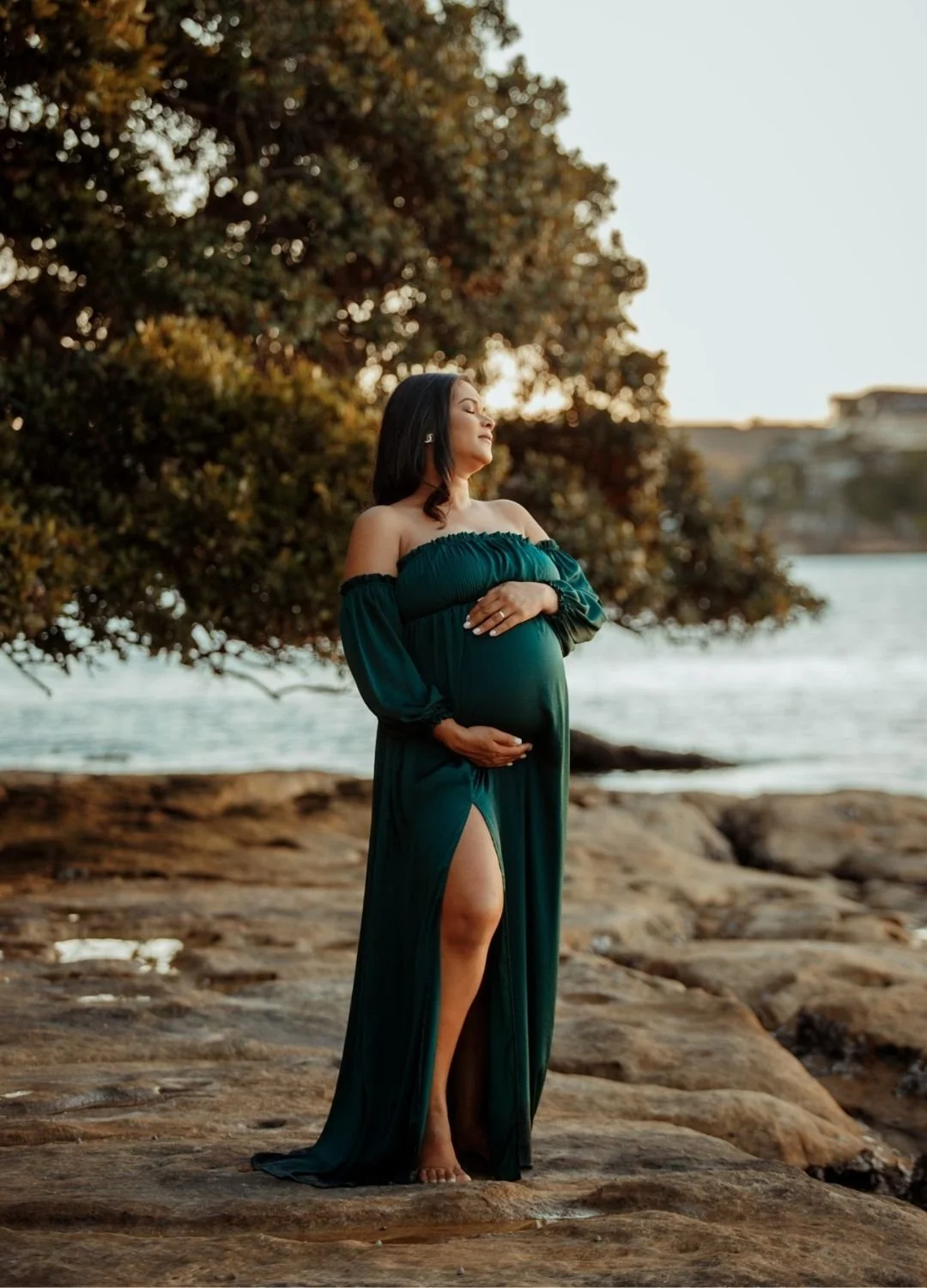 Pregnant woman standing on rocky shore near water, wearing a dark green off-shoulder dress with slit, holding her belly with one hand, eyes closed, peaceful expression, during sunset.