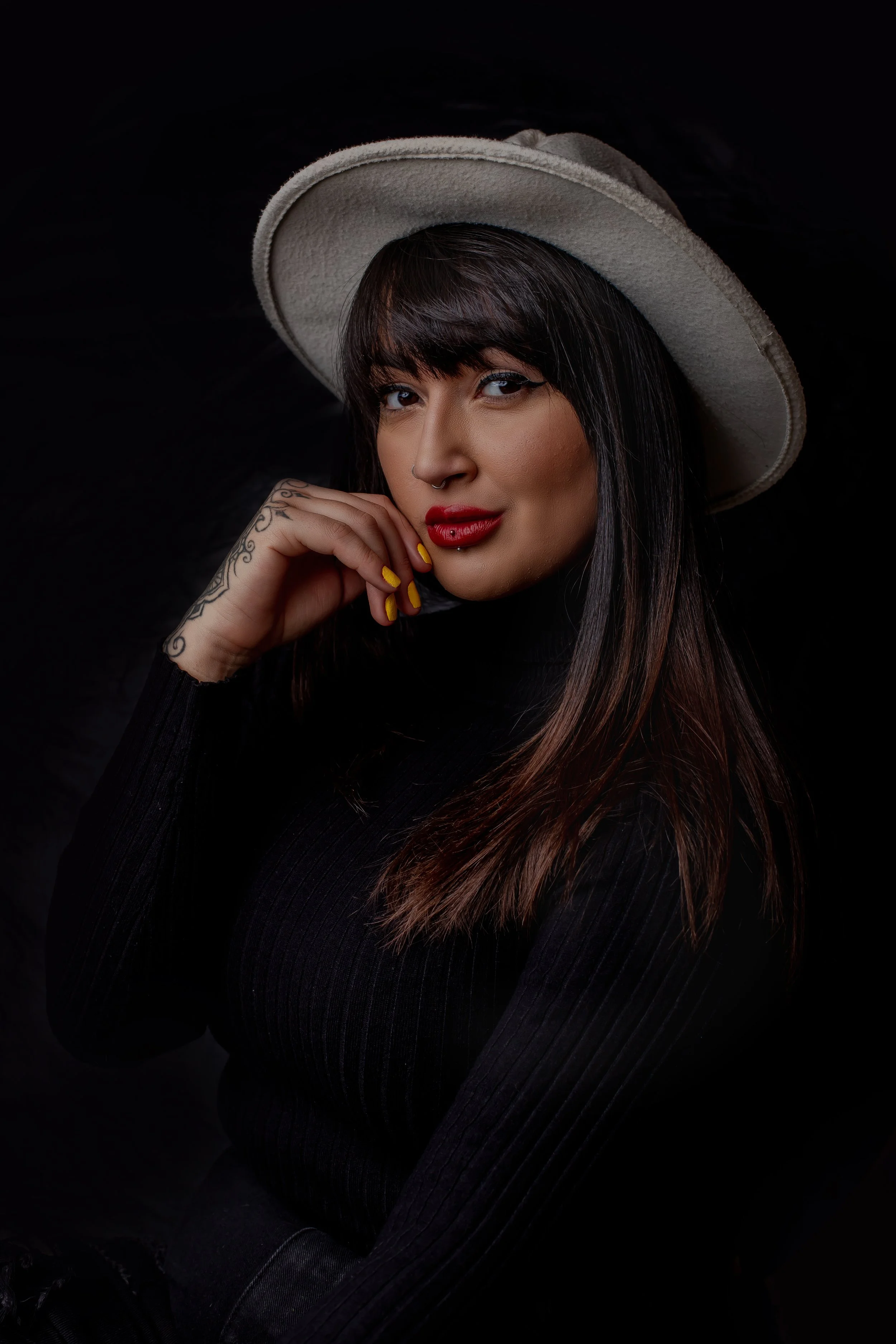 A woman with dark hair, red lipstick, and tattoos on her hand, wearing a beige hat and a black top, posing against a dark background.