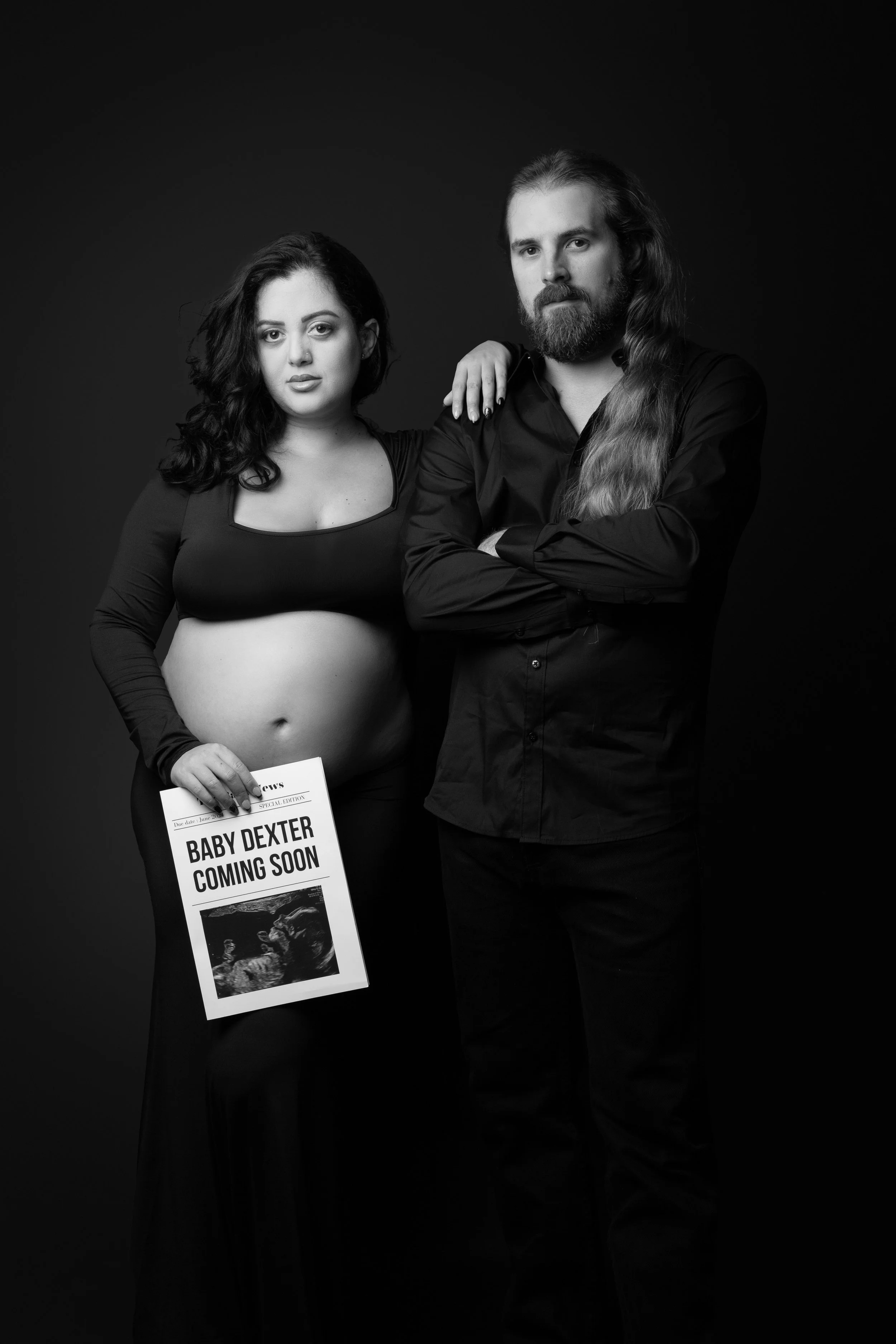 Black-and-white photo of a pregnant woman with dark hair and a man with long hair and beard, standing together against a dark background. The woman is holding a newspaper that says "Baby Dexter Coming Soon" with an ultrasound image. The woman has her