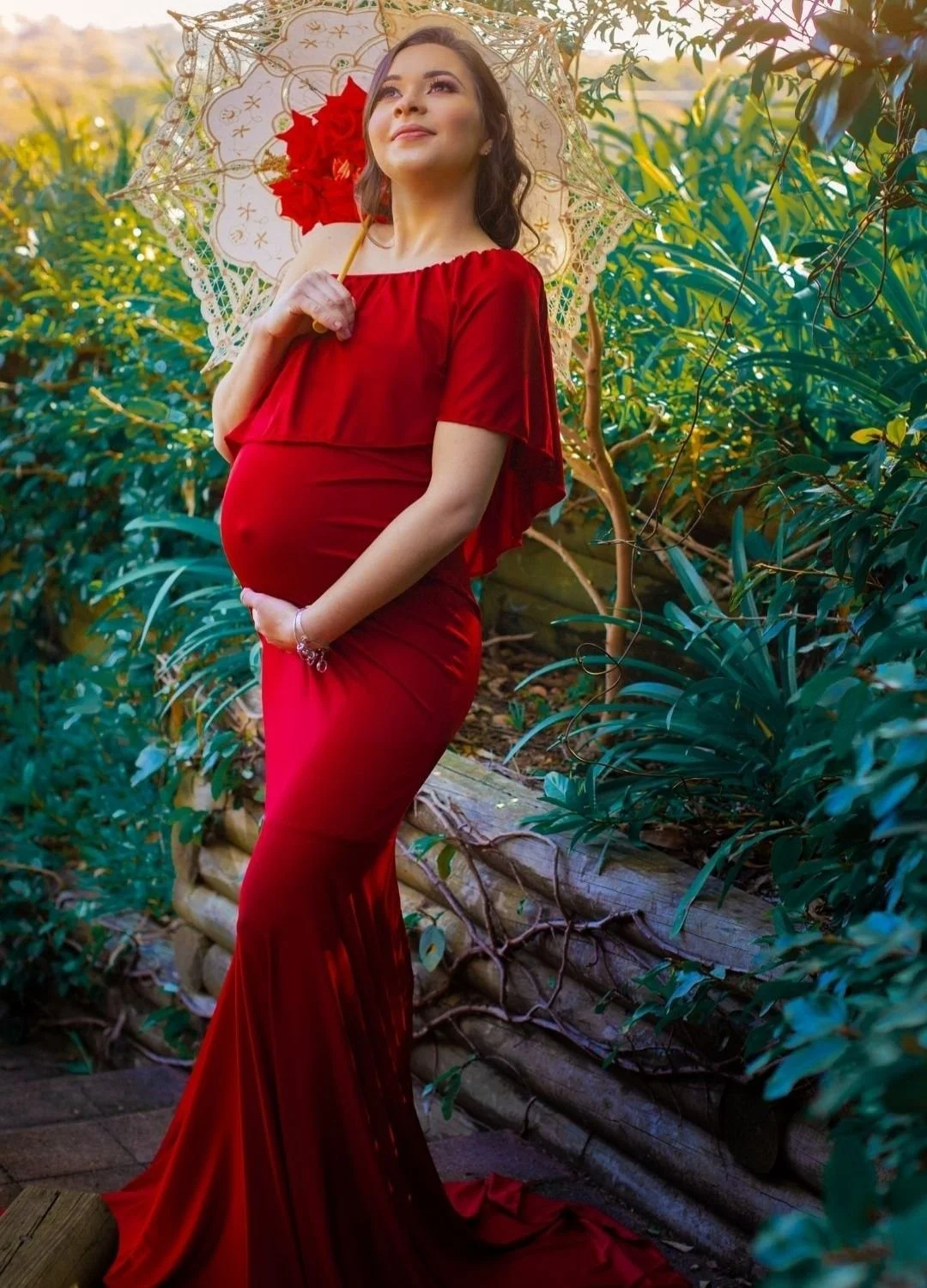 Pregnant woman wearing a red dress holding a red flower parasol in a garden.