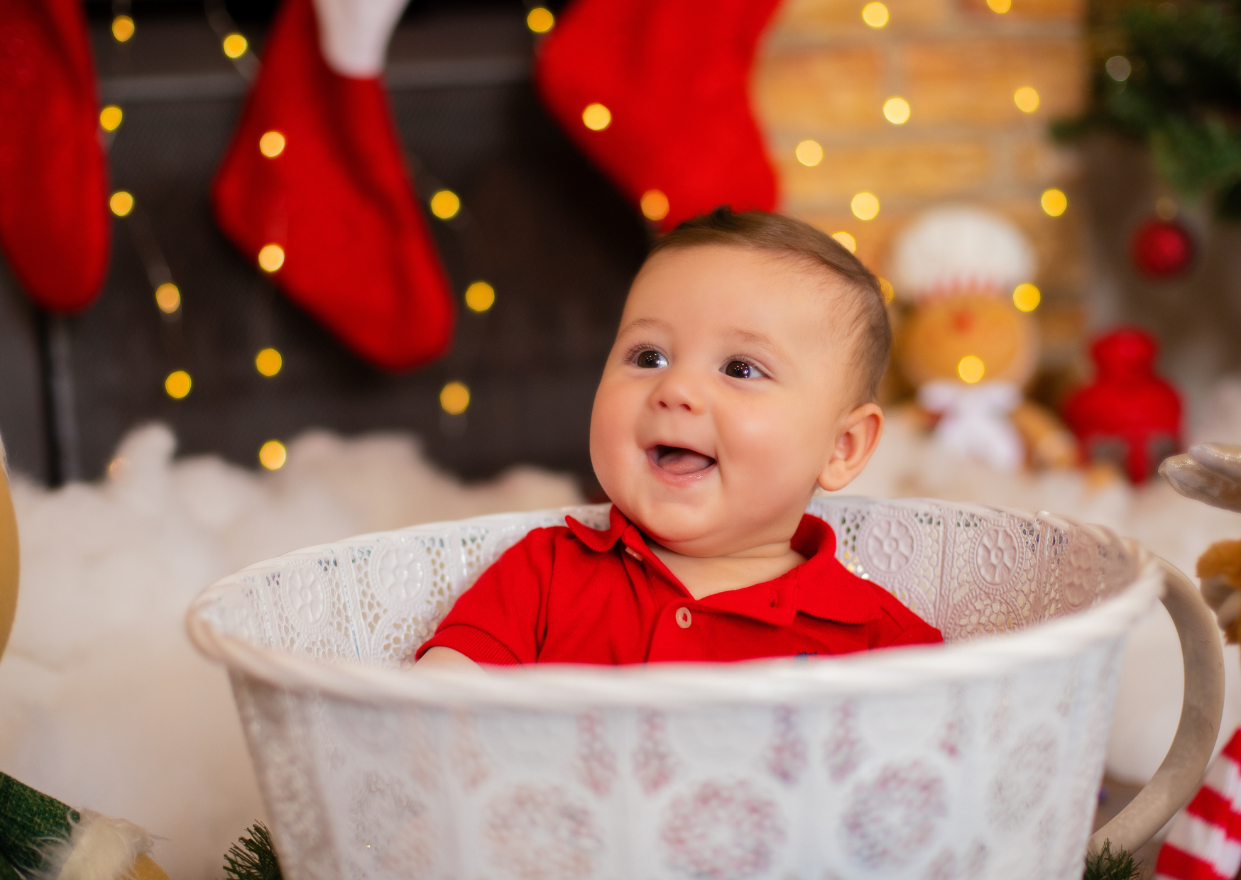 A smiling baby in a red shirt sitting in a white decorative basket. The background features Christmas stockings hanging on the wall, blurred holiday decorations, and a gingerbread man plush toy, creating a festive holiday setting.