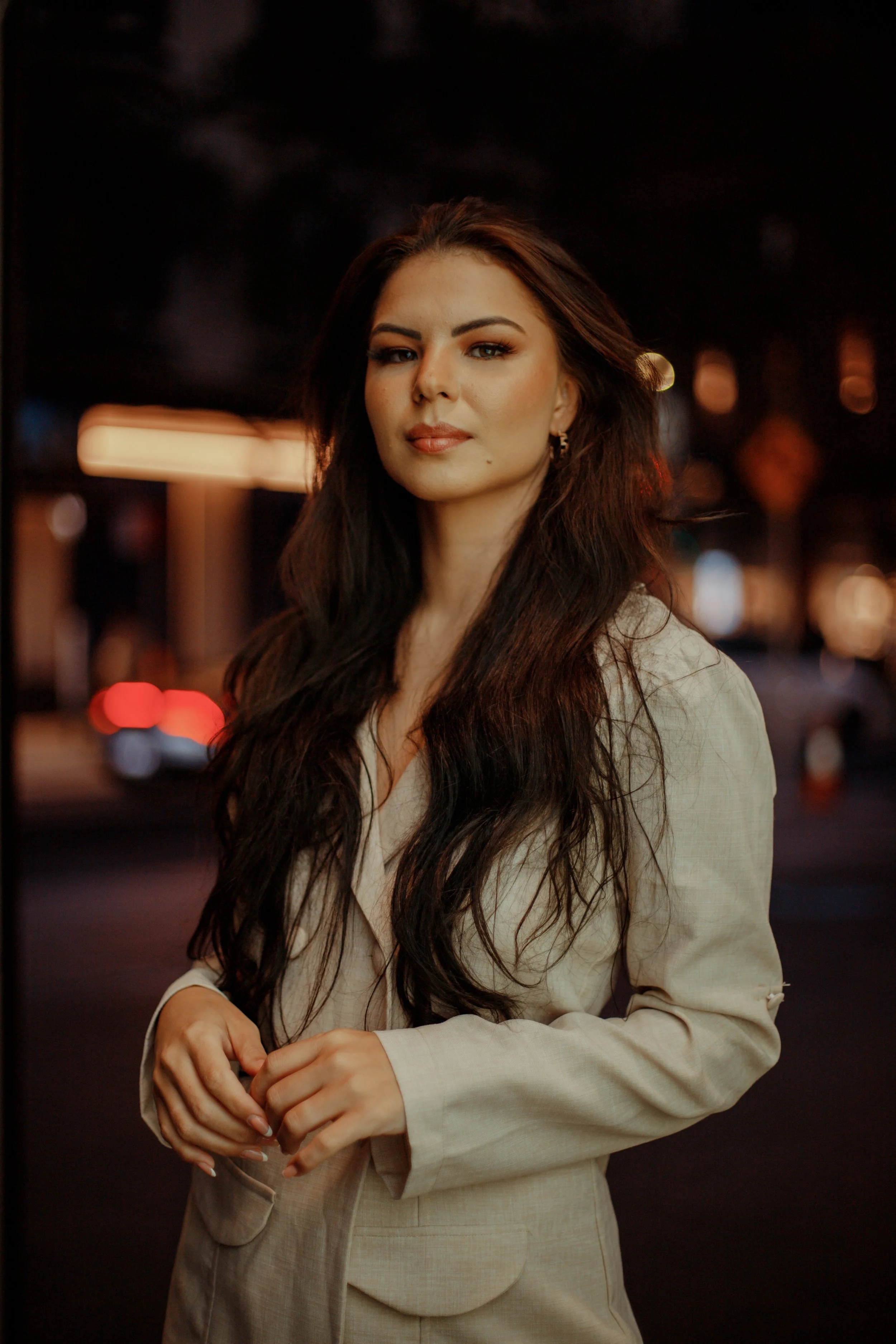A woman with long dark hair and light skin standing outdoors at night, wearing a beige blazer with blurred city lights in the background.