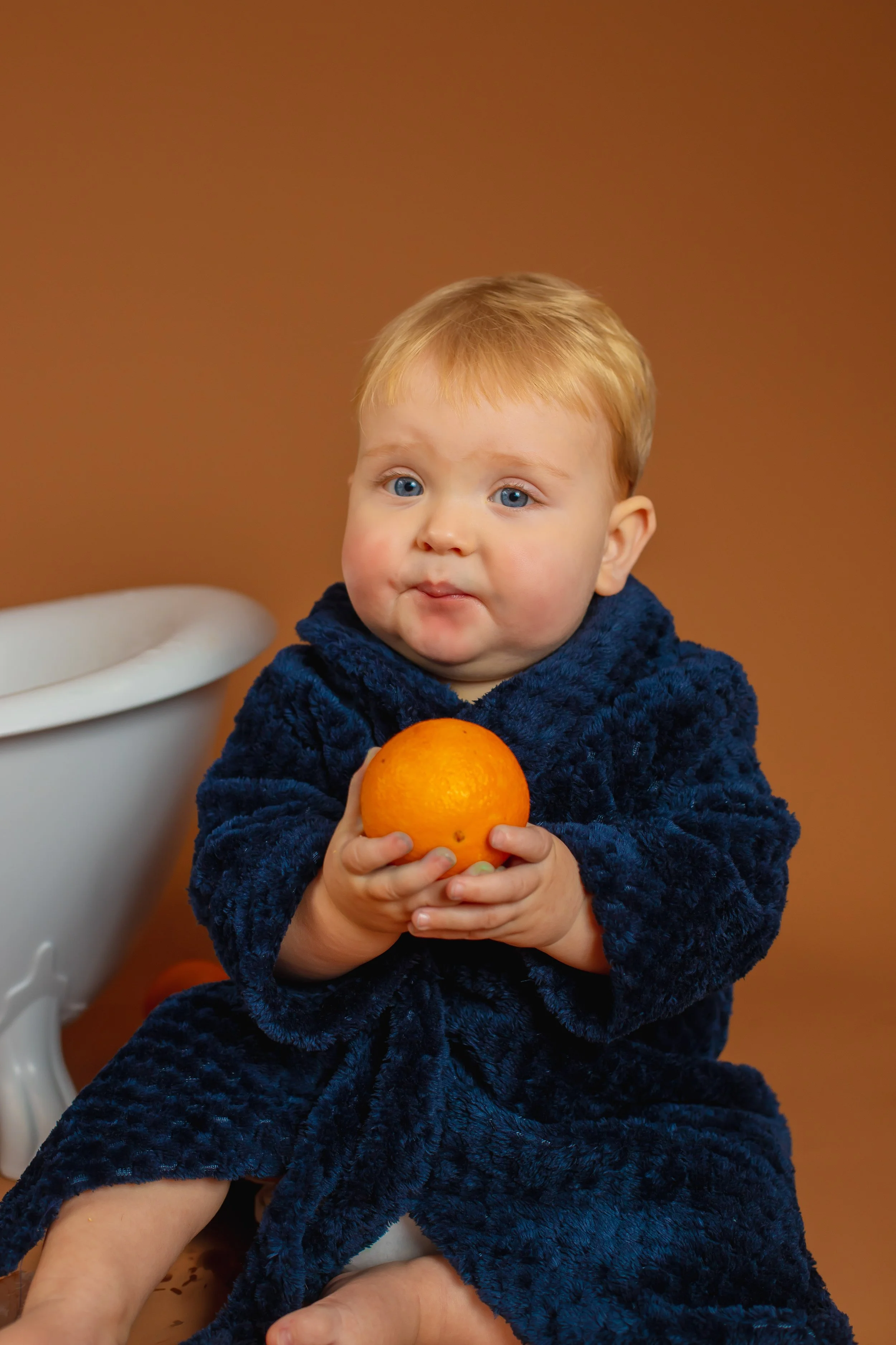 A young child with blue eyes, wearing a navy blue robe, sitting with an orange fruit in their hands against a brown background.