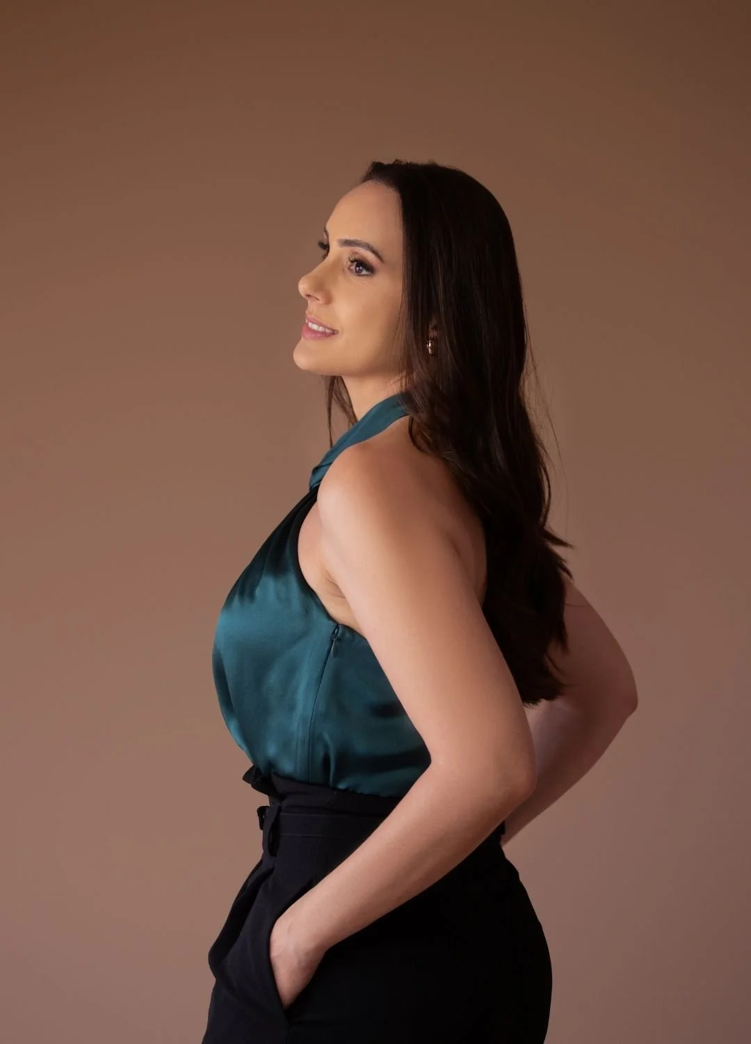 A woman with dark brown hair, wearing a teal satin sleeveless top and black trousers, is standing against a plain background. She is shown in profile with a slight smile, her left hand in her pocket and her right arm relaxed at her side.