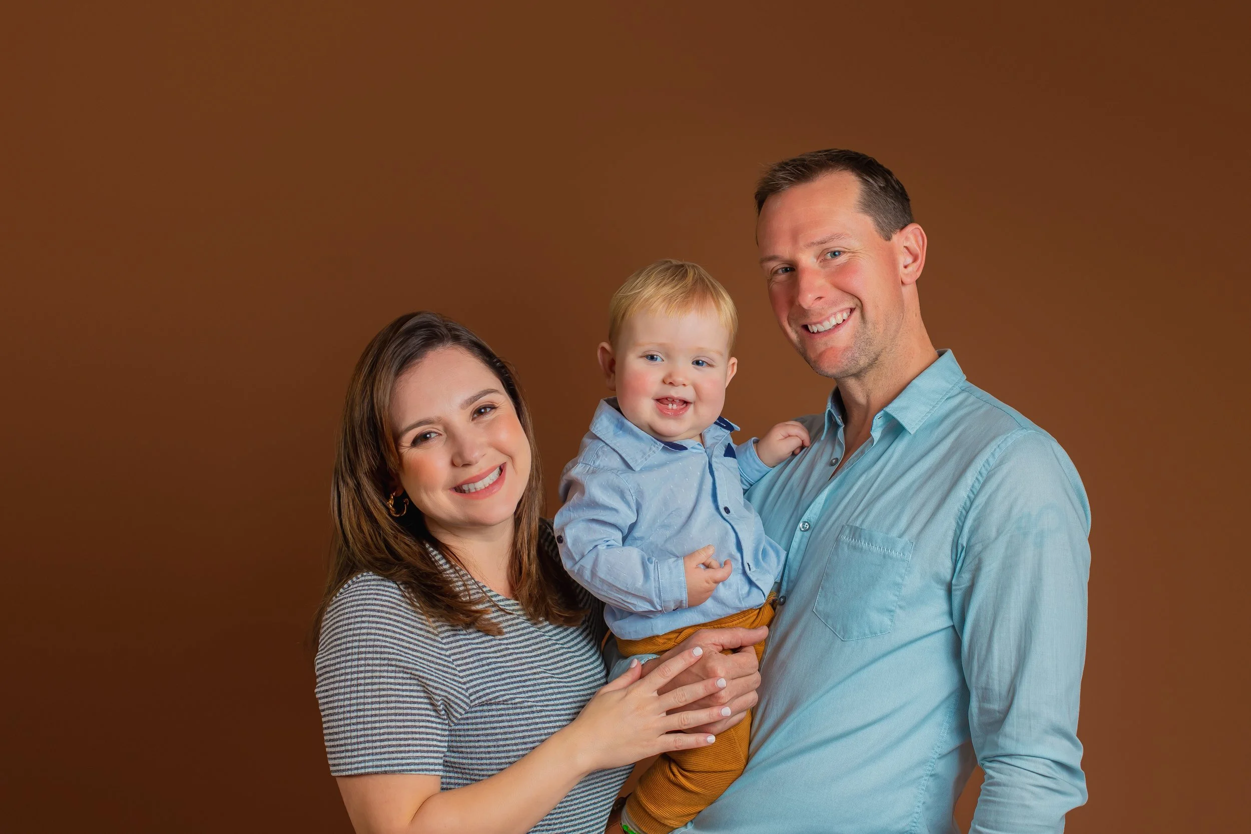 Smiling family of three (mother, father, and young boy) standing together against a brown background, with the father holding the boy.