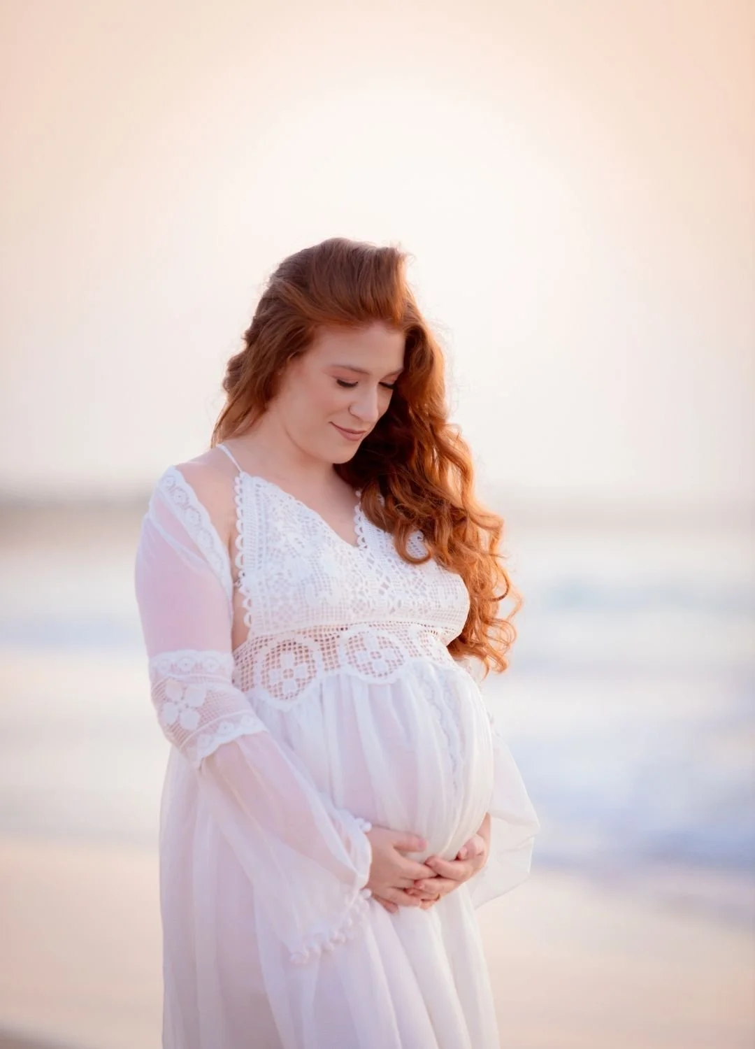 Pregnant woman with long red hair wearing a white lace dress standing on the beach, gently holding her belly and looking down, with a blurred ocean background at sunset.