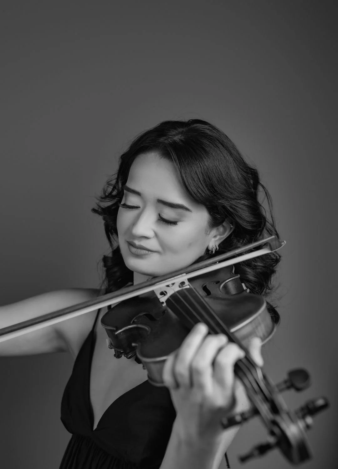 A black and white photo of a woman playing the violin with her eyes closed, wearing earrings and a sleeveless top.