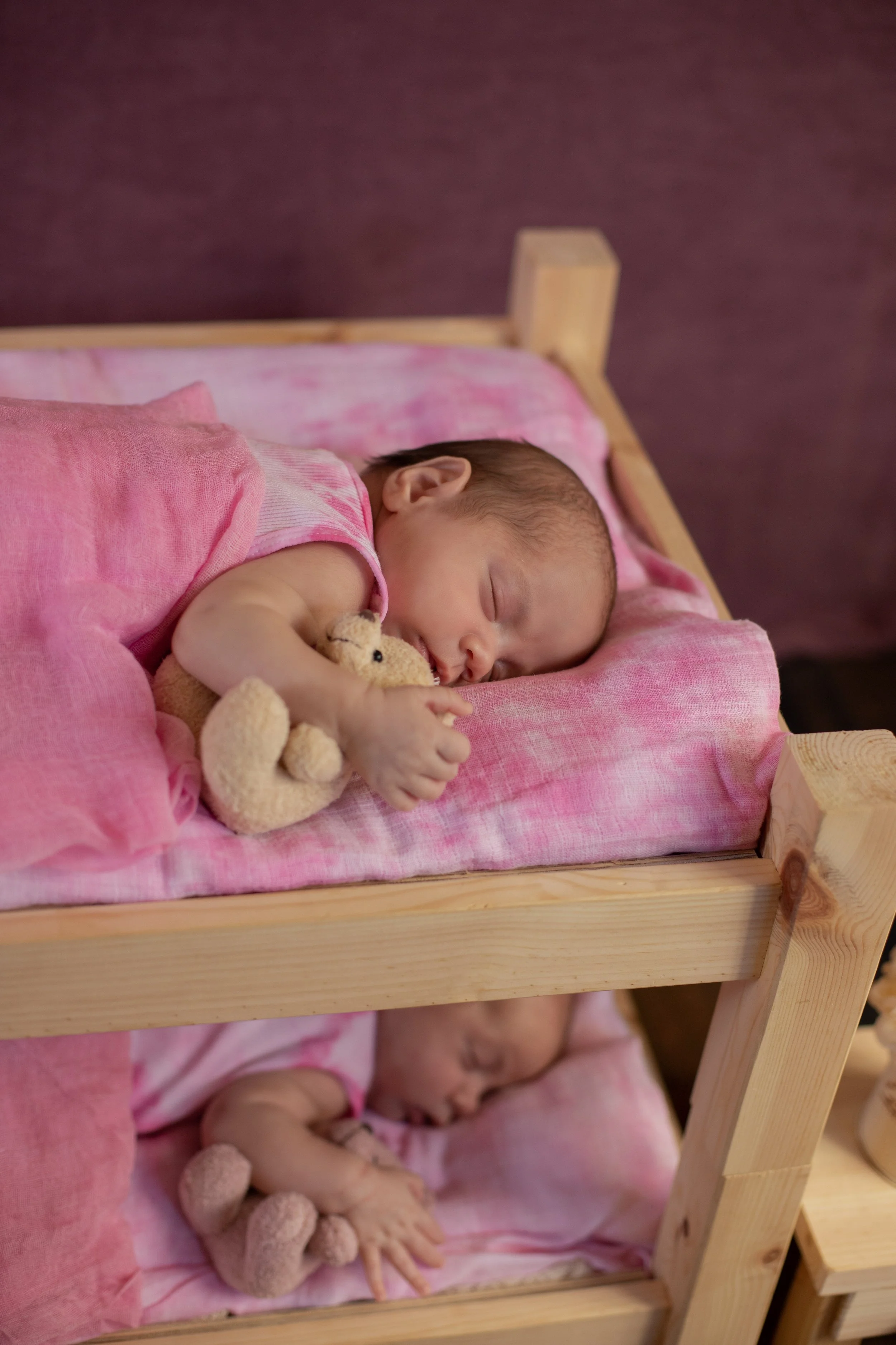 A baby sleeping on a pink bed, hugging a small stuffed teddy bear, with another baby sleeping on a lower bed in the background.