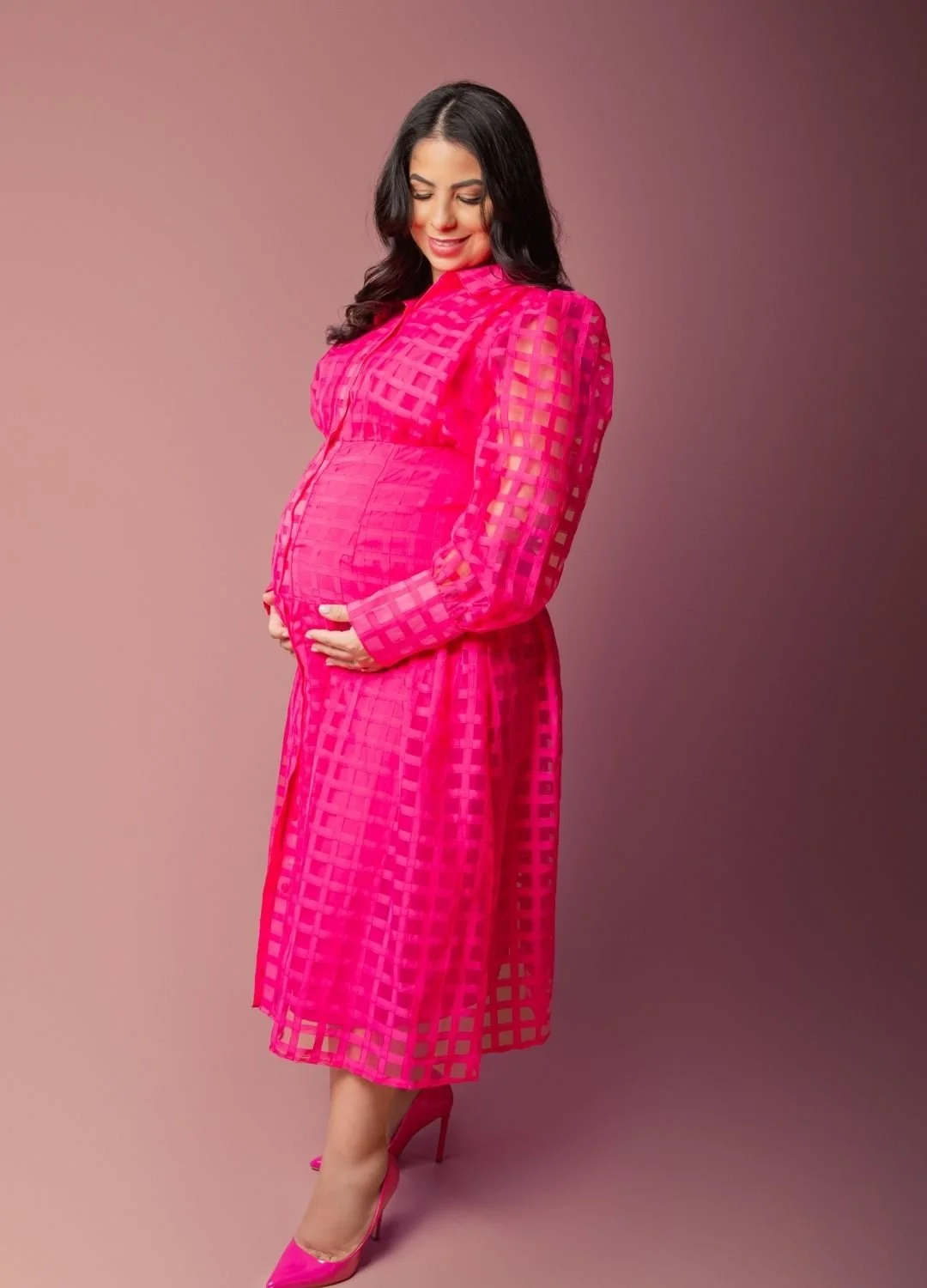 A pregnant woman in a bright pink, semi-transparent, checkered dress with matching high heels, standing against a gradient pink background, smiling while holding her belly.