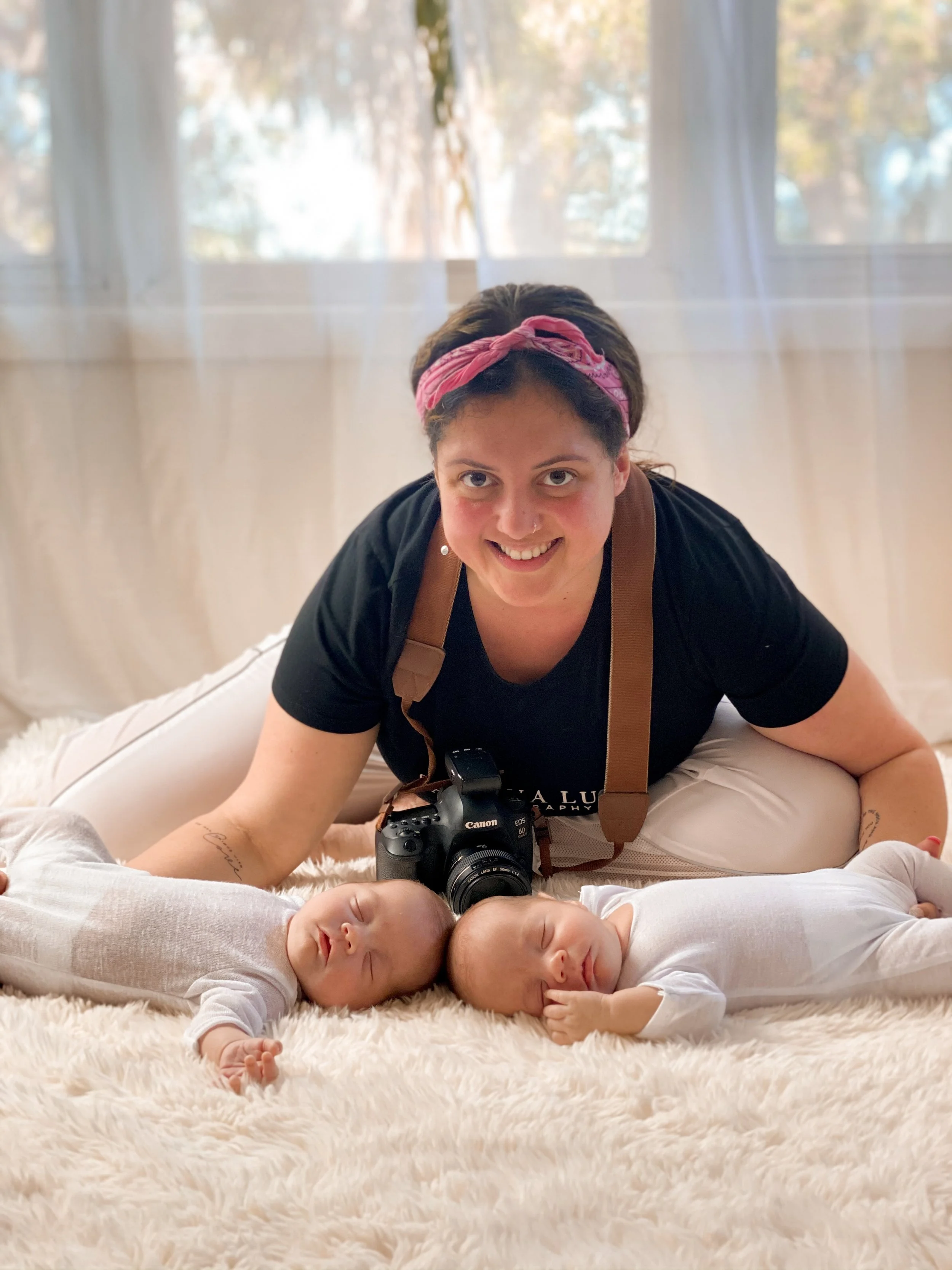 A woman with a pink headband and black shirt, smiling and leaning over two sleeping babies on a fluffy white rug near a window.