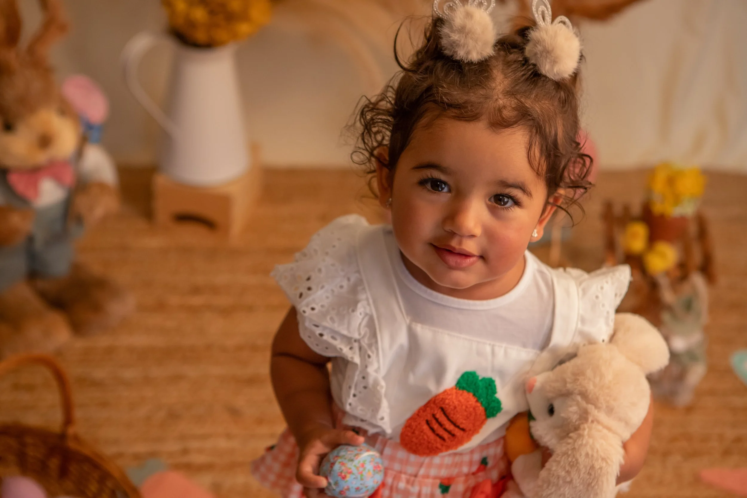 Young girl with curly hair wearing a white dress with eyelet sleeves, holding a stuffed teddy bear and a small decorative ball, smiling indoors with stuffed animals and decorations in the background.