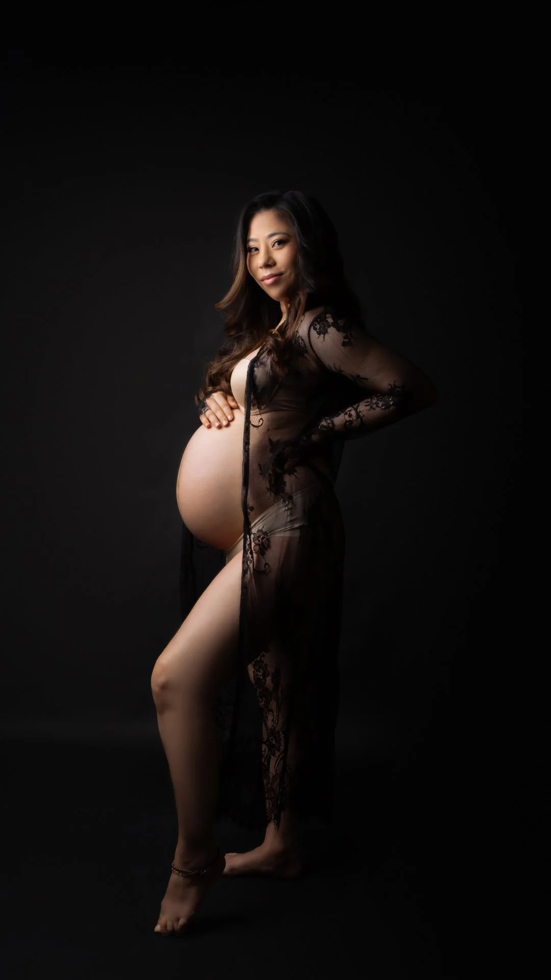 A pregnant woman in a sheer black lace dress against a black background, standing with one hand on her belly and the other on her hip, looking confidently at the camera.
