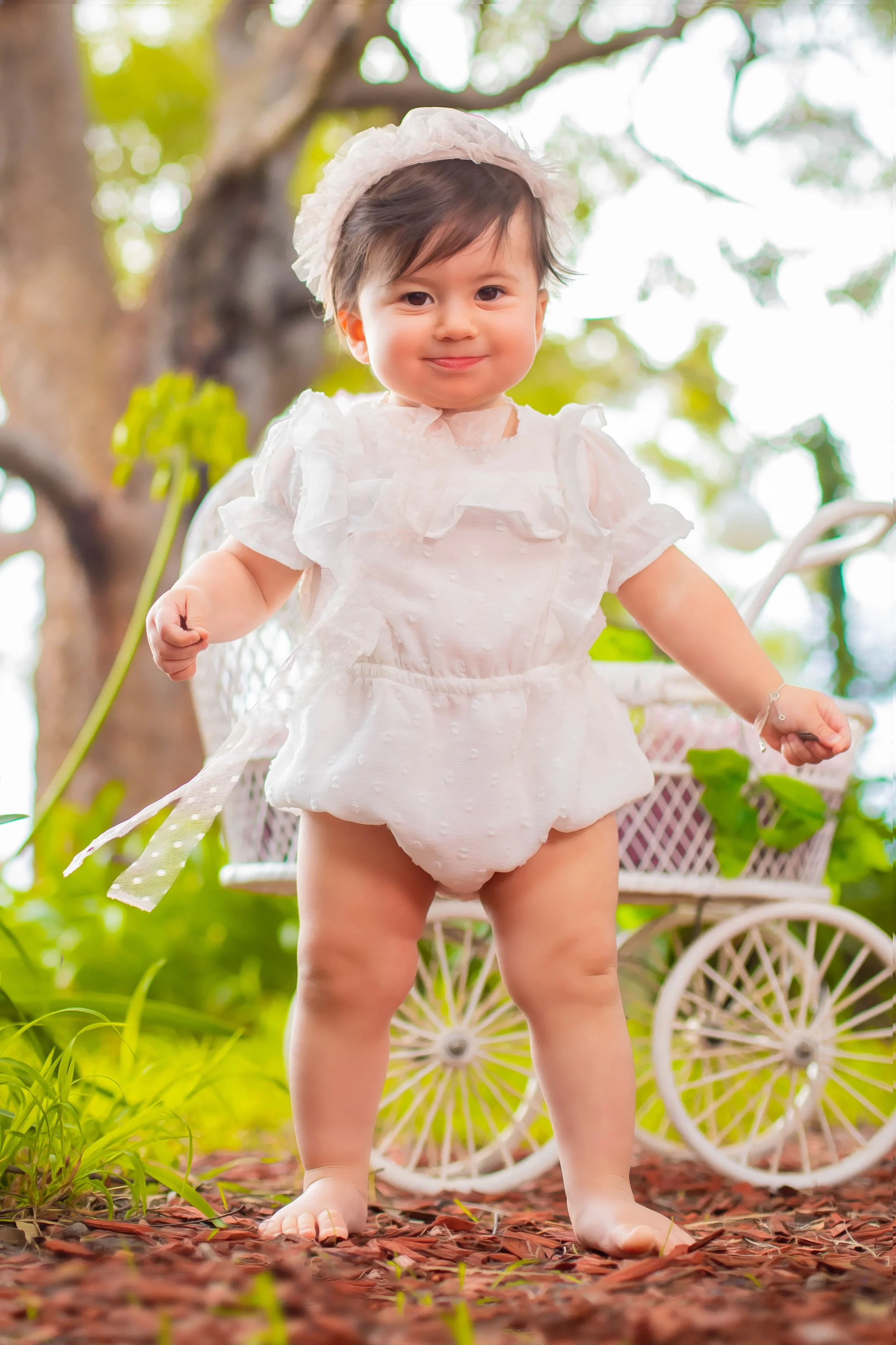 A young toddler girl dressed in a white outfit with a matching headband, standing outdoors on a bed of red mulch with a white decorative baby carriage behind her. The background features green foliage and a large tree.