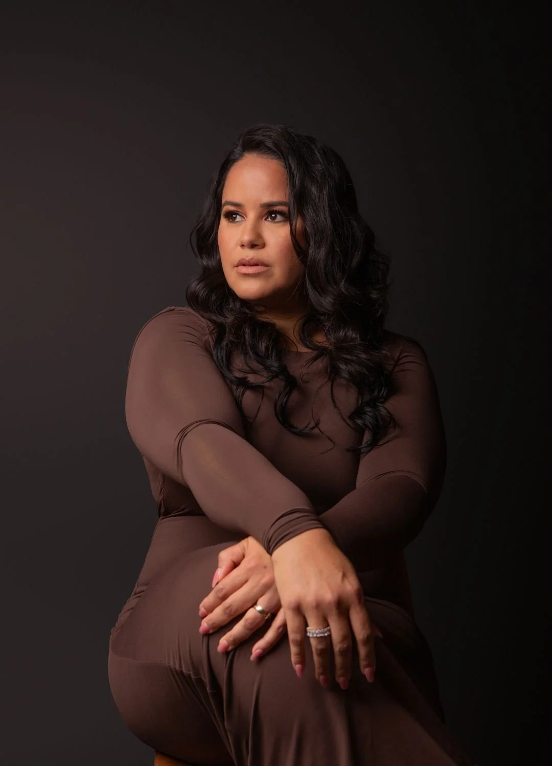 A woman with dark, wavy hair and makeup, wearing a sheer brown dress, sitting against a dark background, looking to the side.