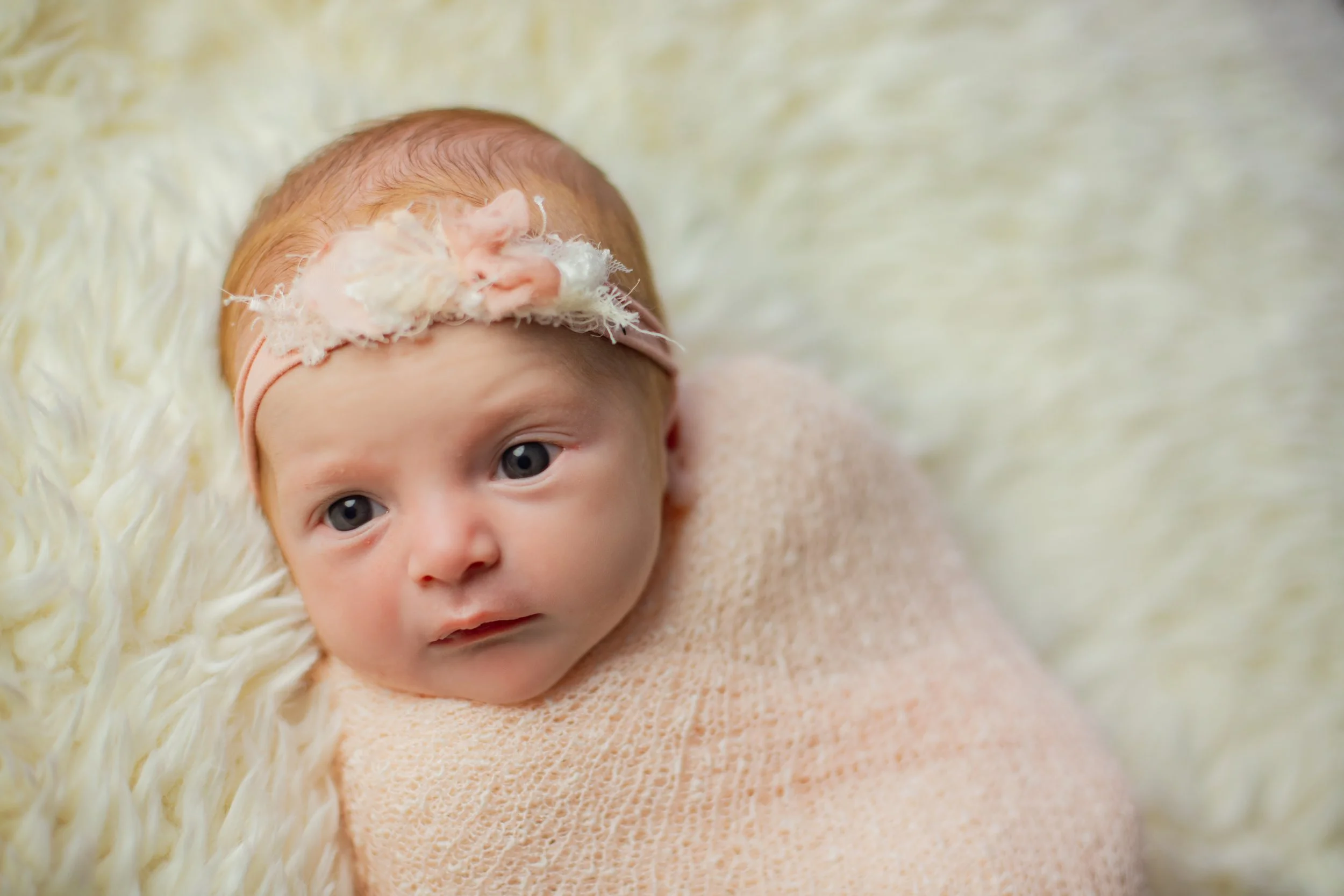 Close-up of a newborn baby with blue eyes, wearing a pink headband with a floral decoration and wrapped in a soft pink blanket, lying on a fluffy cream-colored surface.