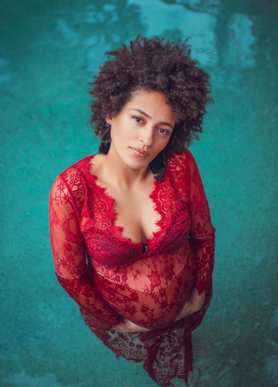 A woman with curly hair standing in a pool, wearing a red lace dress.