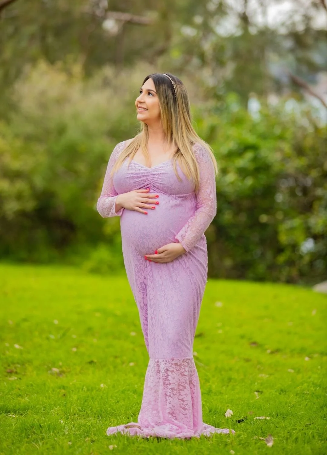 Pregnant woman in a pink lace dress standing on green grass outdoors, smiling and cradling her belly.