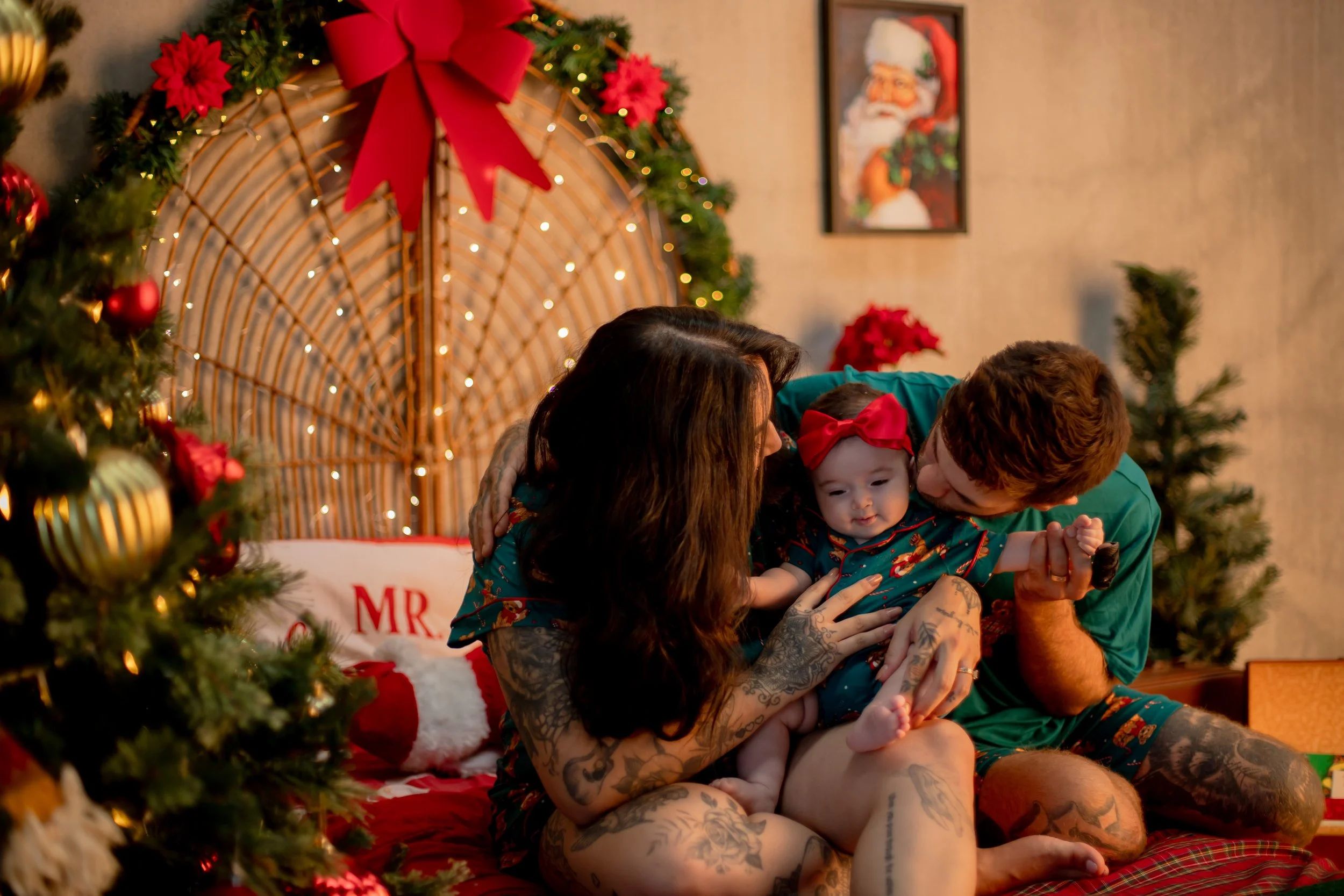 A family sitting on a red bed in a Christmas decorated room, with a Christmas tree on the left and festive decorations, including a large red bow and poinsettias, behind them. The family includes a woman with tattoos, a man, and a baby girl wearing a