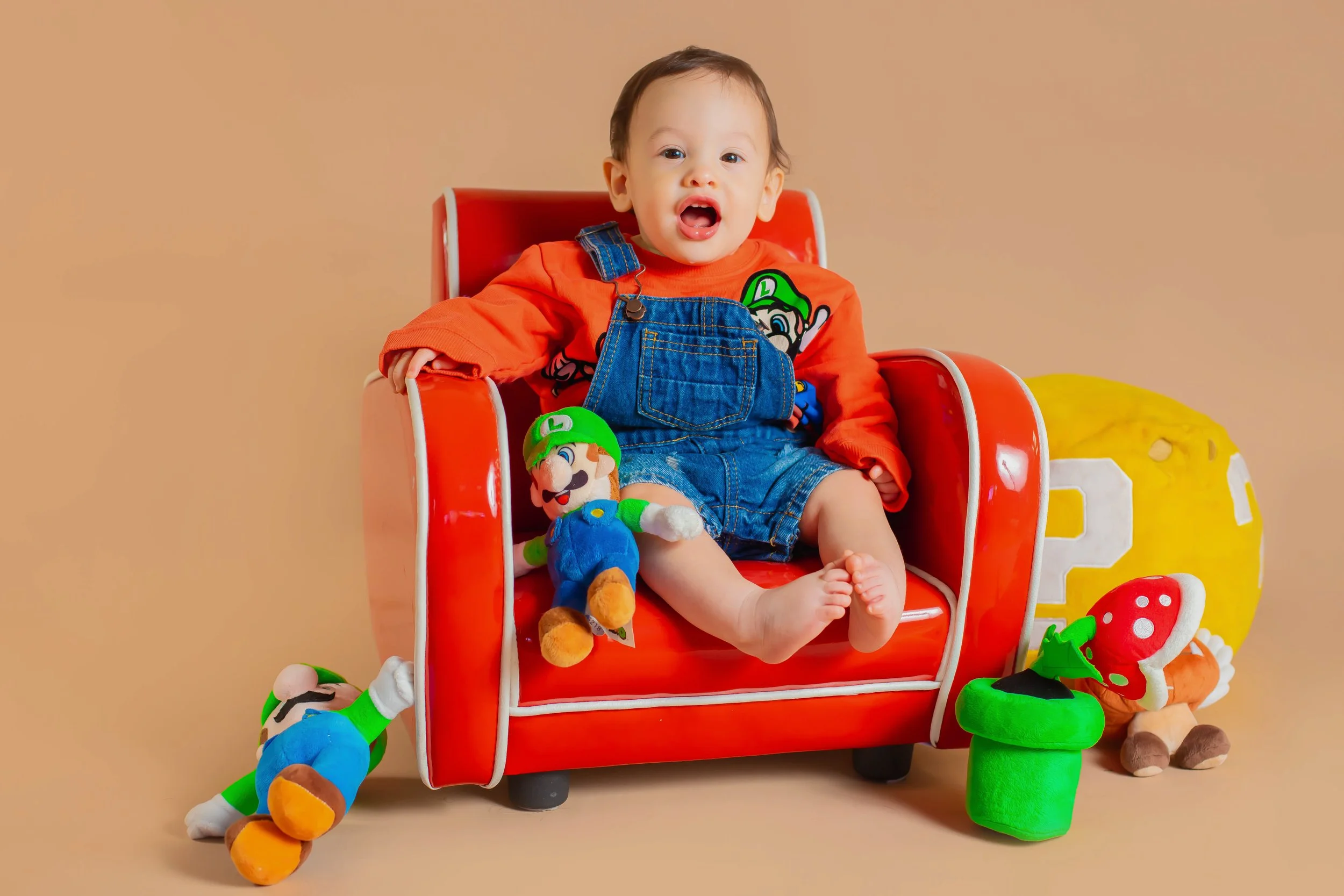 Child sitting in a red chair surrounded by Mario themed plush toys, including a Mario plush on the child's lap and Luigi plush on the floor, with a yellow question mark block and a red mushroom plush nearby.