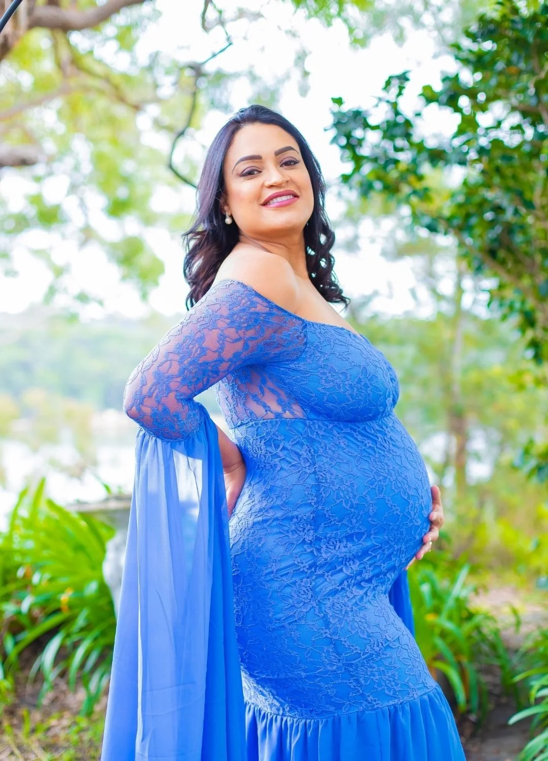 Pregnant woman outdoors in a blue lace off-shoulder dress, smiling, with trees and greenery in the background.