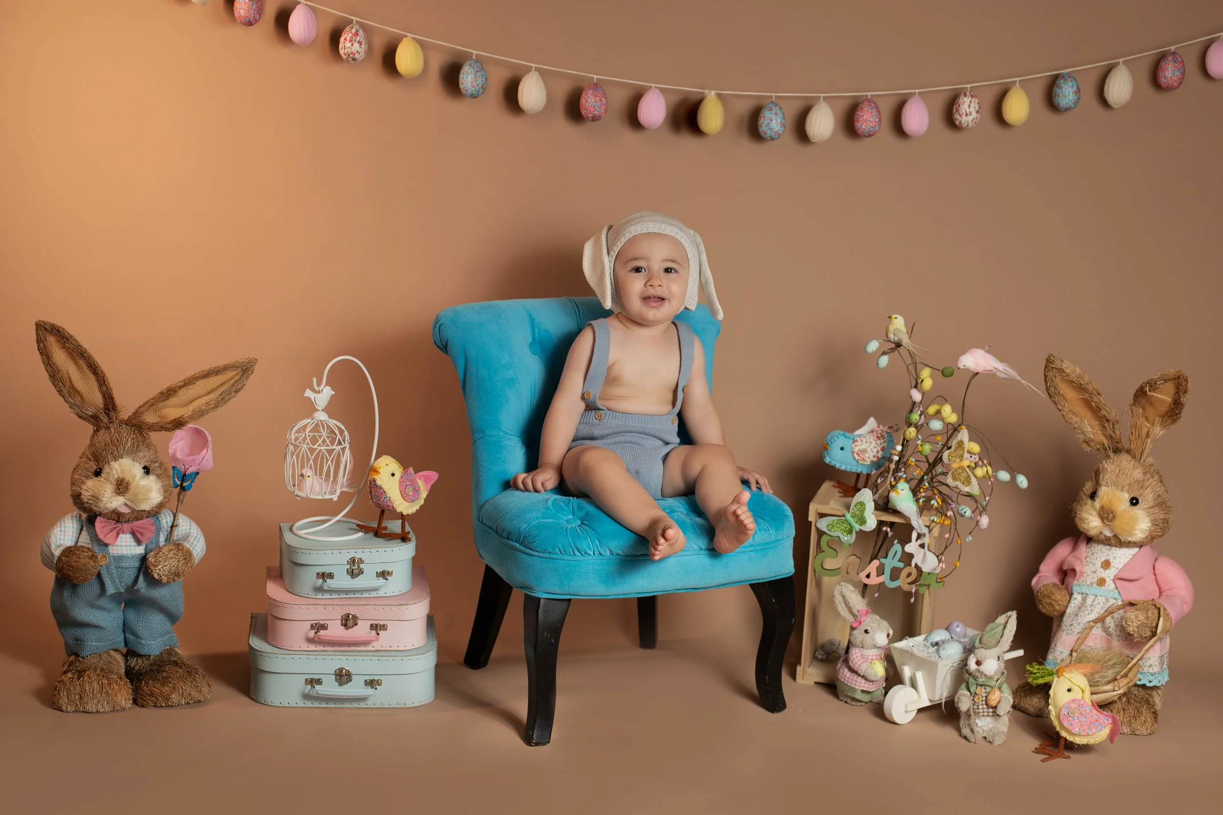 Child sitting in a blue chair during Easter celebration, surrounded by plush bunny stuffed animals, decorative eggs, small chairs with rabbits and eggs, through a Christmas style garland and Easter-themed decorations.