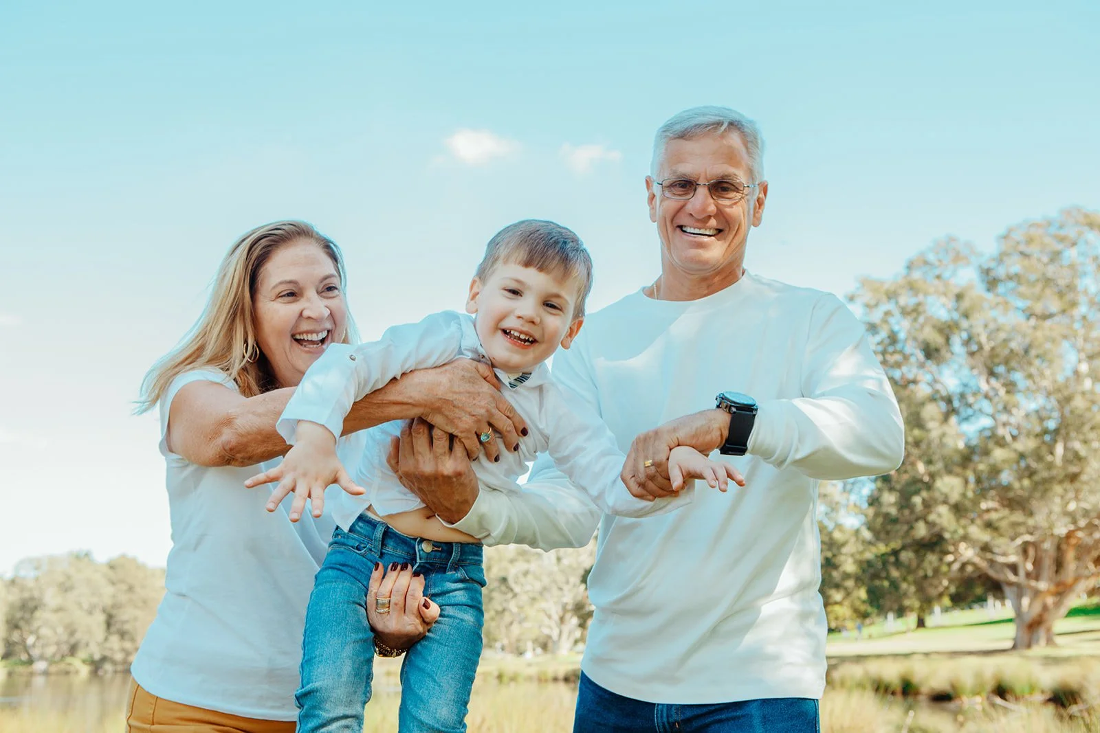 Smiling elderly couple holding young boy outdoors in a park on a sunny day.