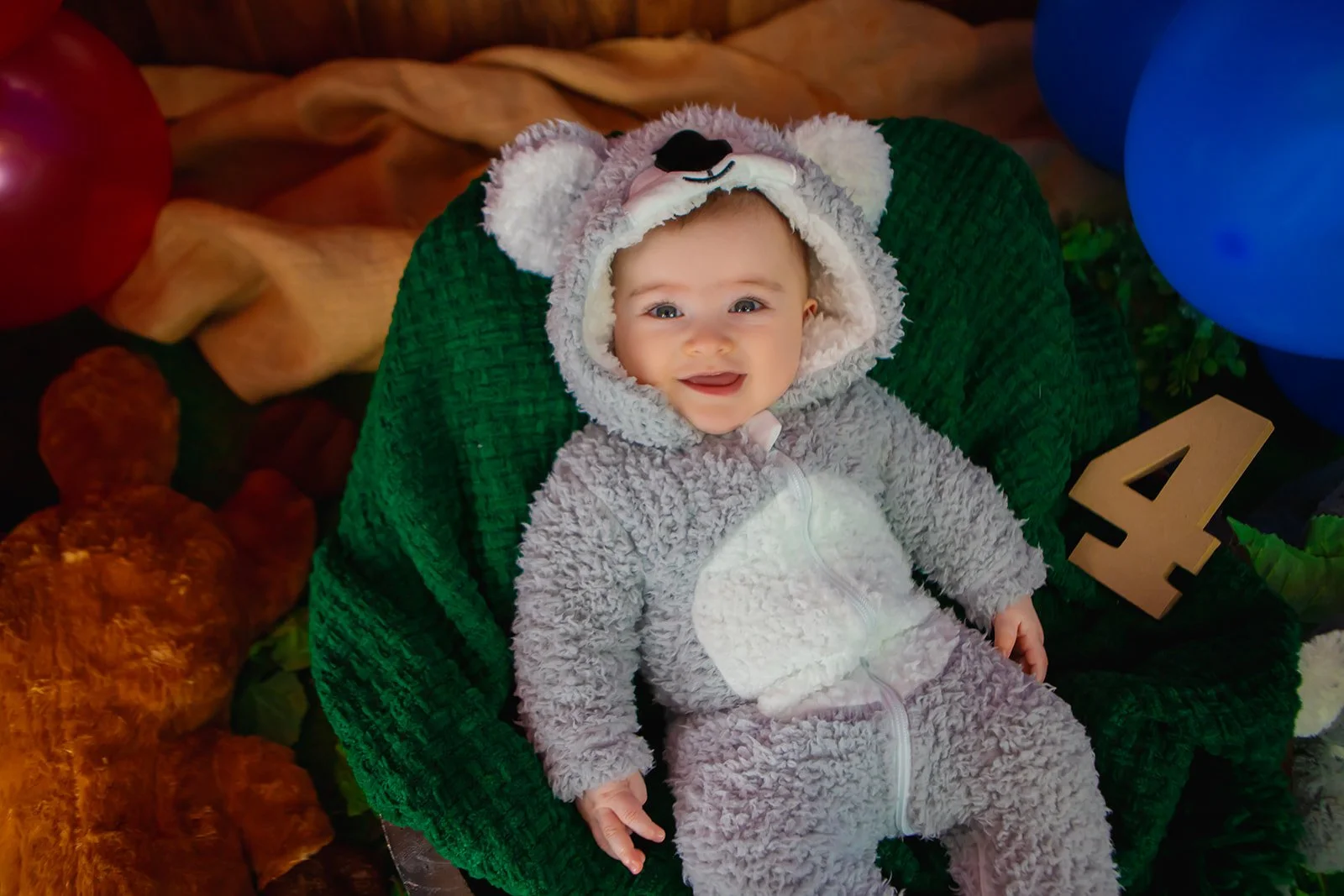 Smiling baby in a bear costume sitting in a green chair with a wooden carved number 4 next to them, surrounded by colorful balloons and blankets.