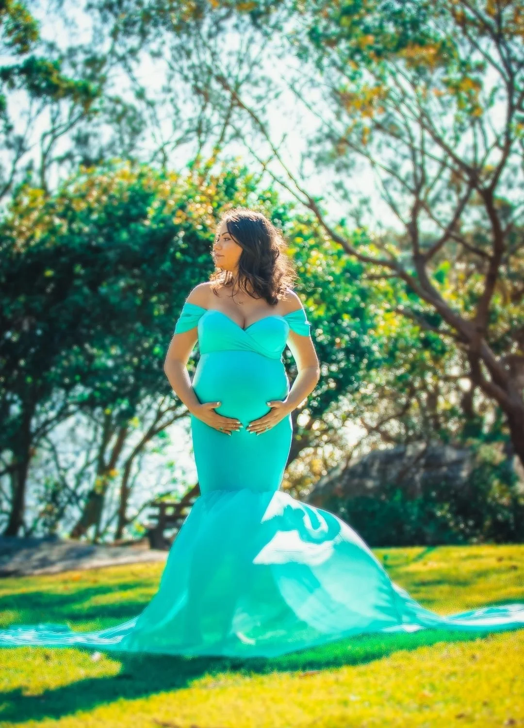 A pregnant woman in a turquoise dress standing outdoors in a park with green trees in the background.