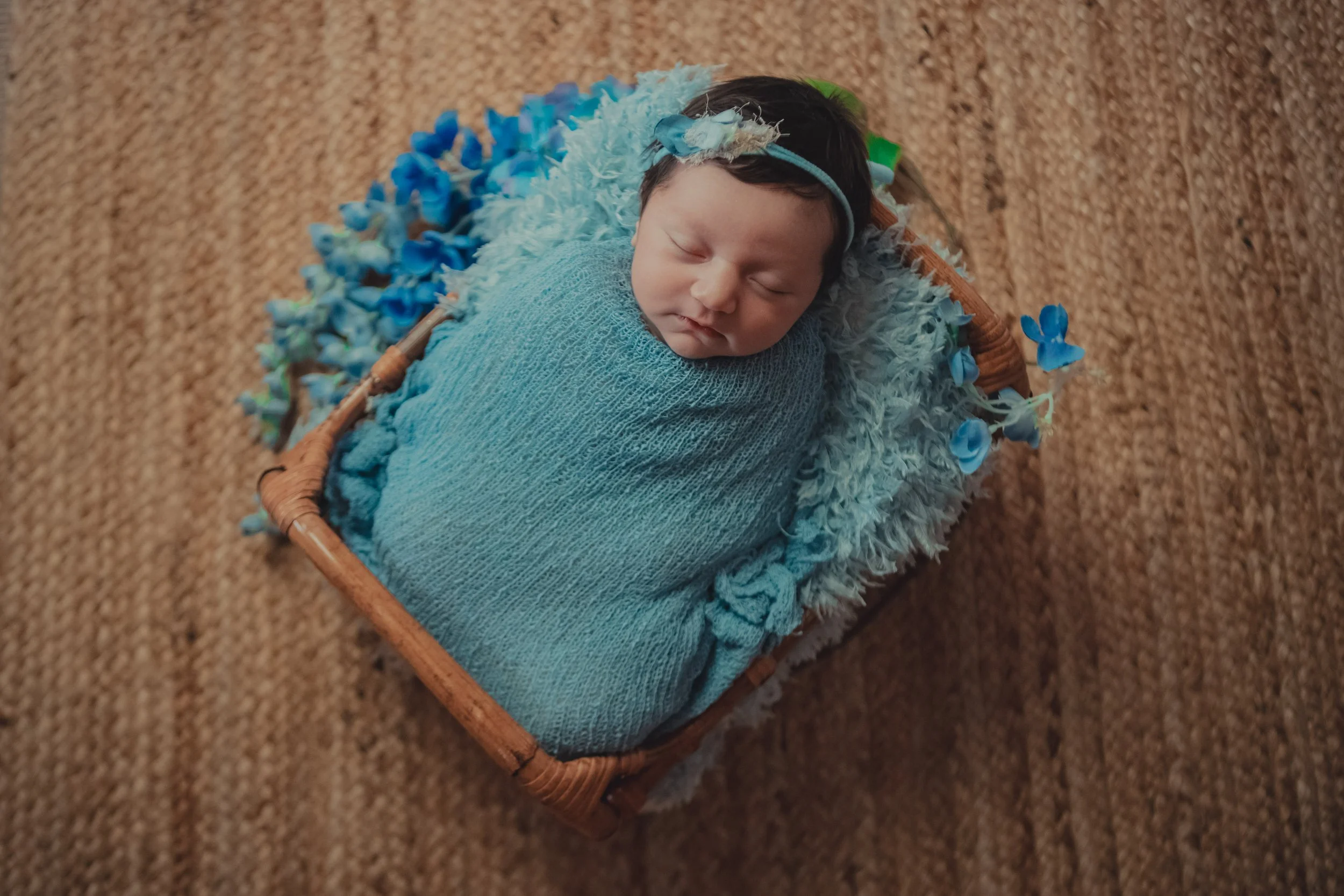 A newborn baby swaddled in a light blue blanket, sleeping in a wicker basket lined with soft feathers and surrounded by blue artificial flowers, on a brown textured surface.
