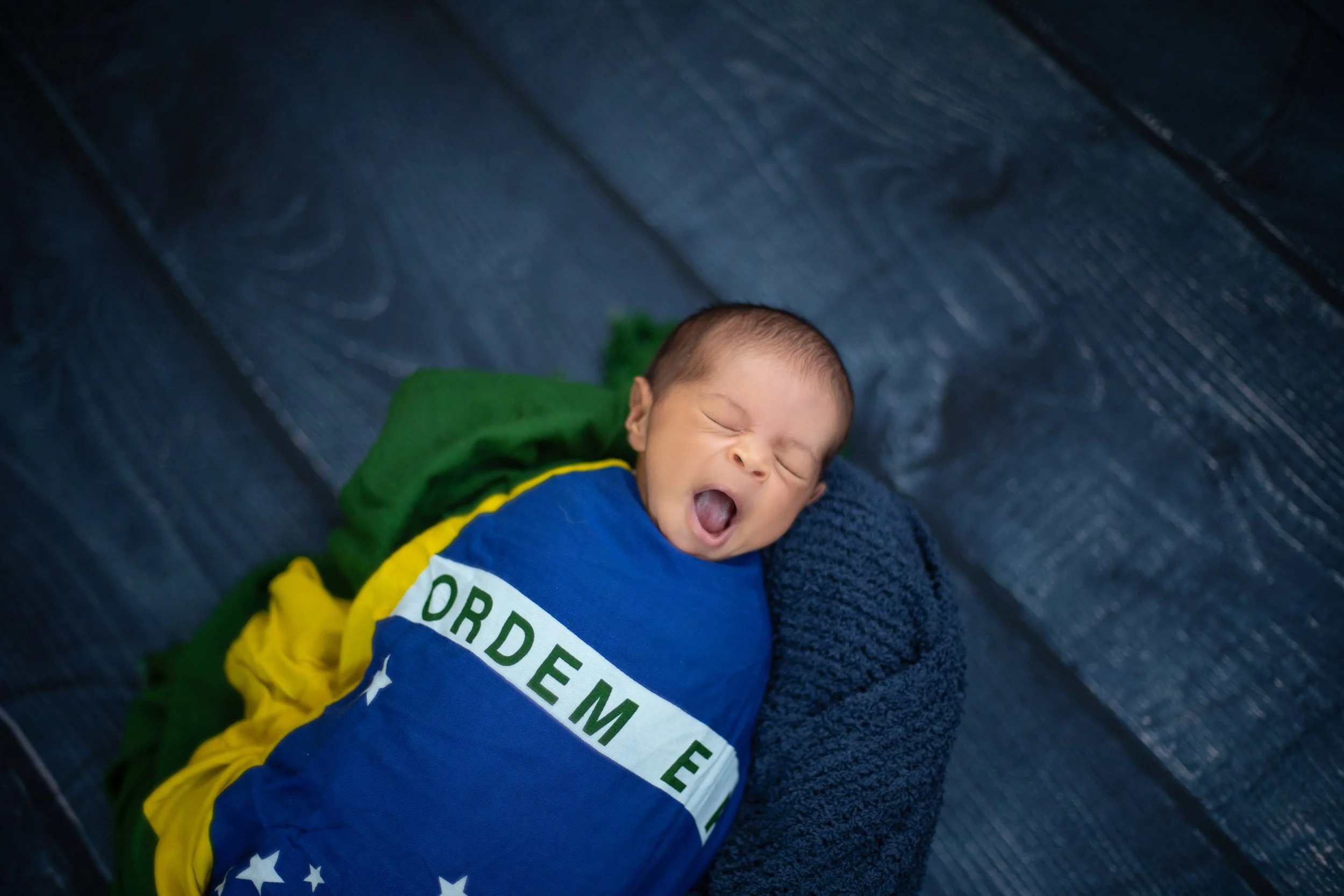 A yawning newborn baby lying on a dark textured surface, wrapped in a rainbow-themed blanket with the words 'ORDER EM' and stars, with a green and yellow cloth nearby.